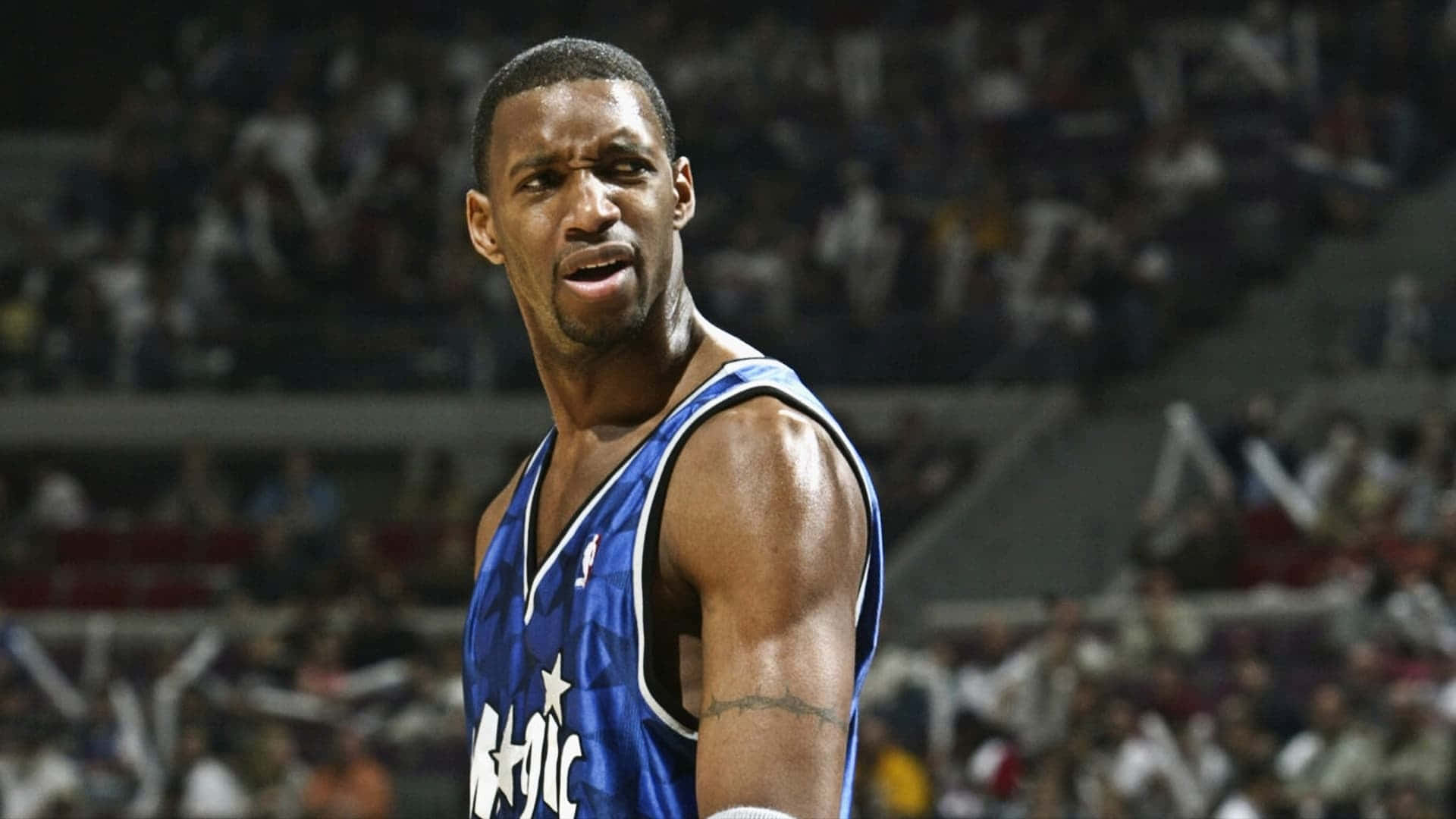Tracy Mcgrady 桌布 壁紙 — 一位專注的籃球運動員身穿藍色奧蘭多魔術隊球衣，站在球場上，背景是模糊的人群。
