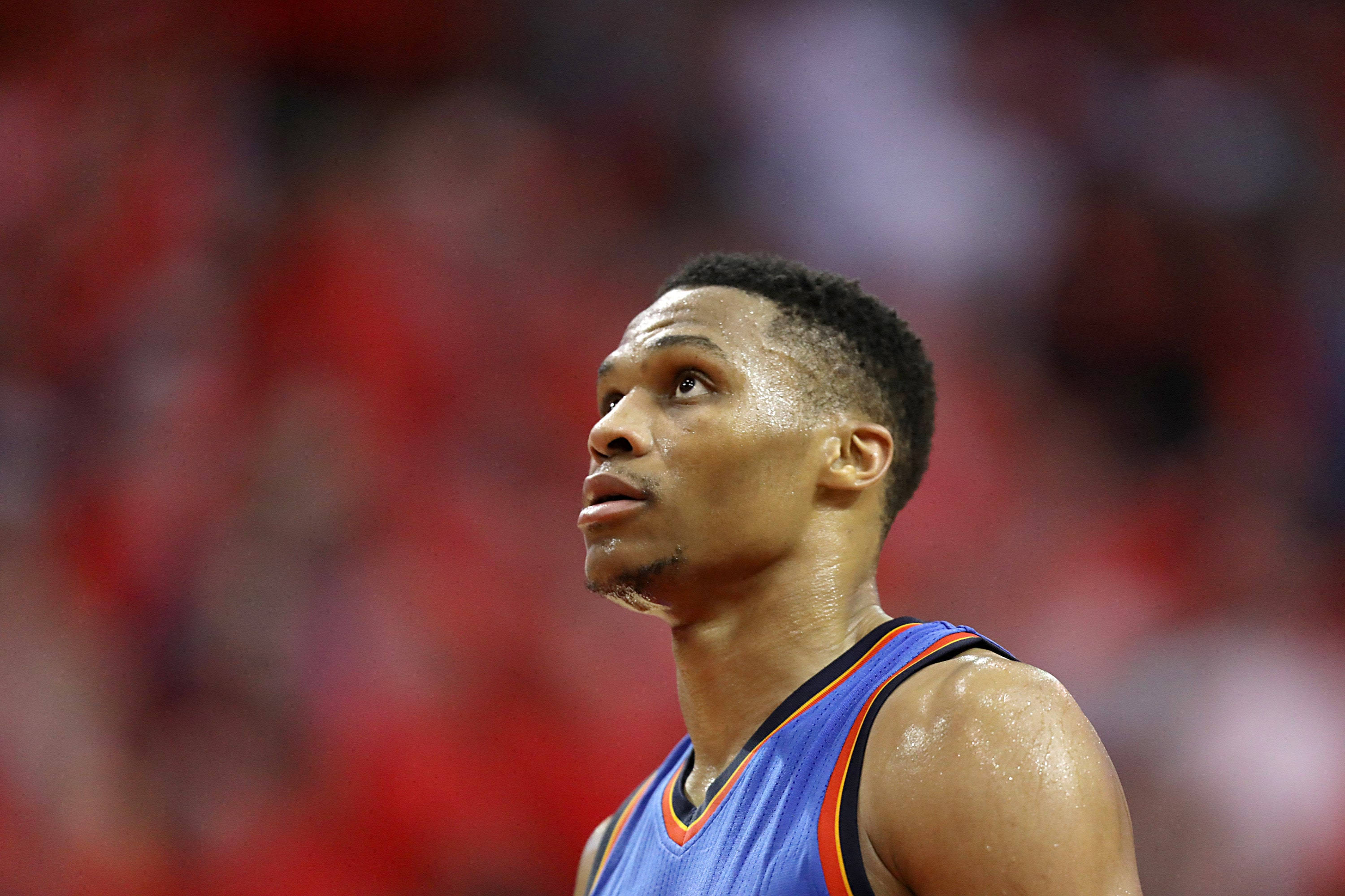 Westbrook 桌布 壁紙 — 一位專注的籃球運動員身穿藍色球衣，在模糊的紅色人群背景下堅定地向上看。