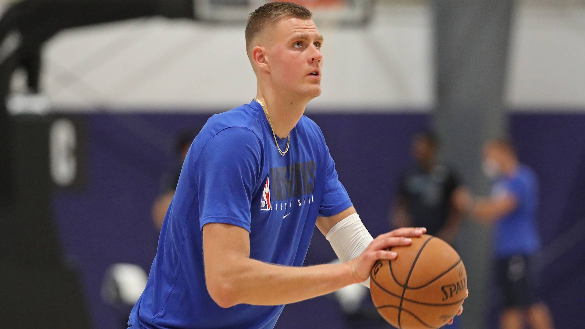 Kristaps Porzingis 桌布 壁紙 — 一位專注的籃球運動員身穿藍色球衣，在室內球場訓練中持球，背景有模糊的隊友。