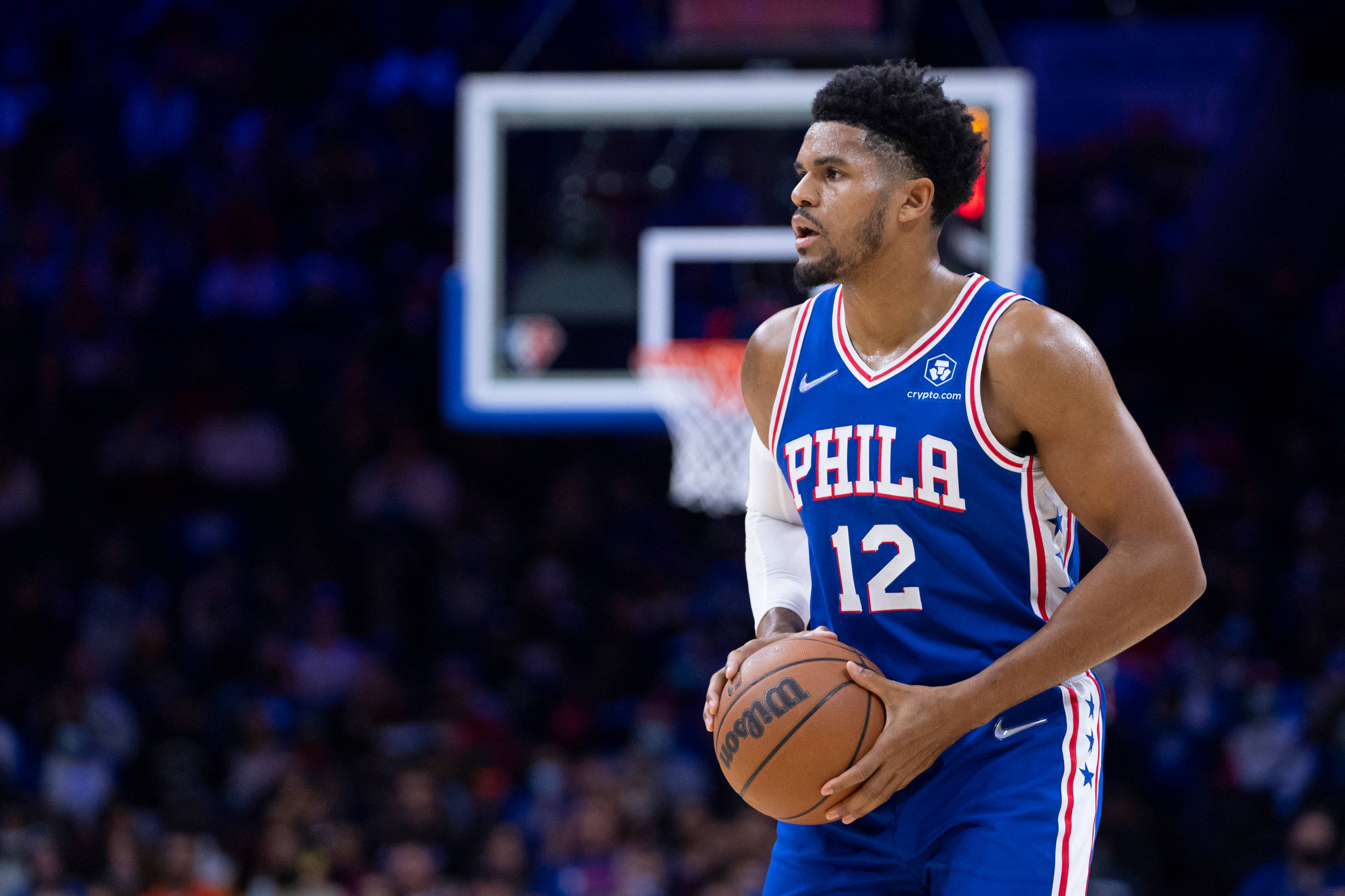 76ers 桌布 壁紙 — 一位專注的籃球運動員身穿藍色費城球衣，在場館燈光下持球，身後是模糊的人群和籃框。