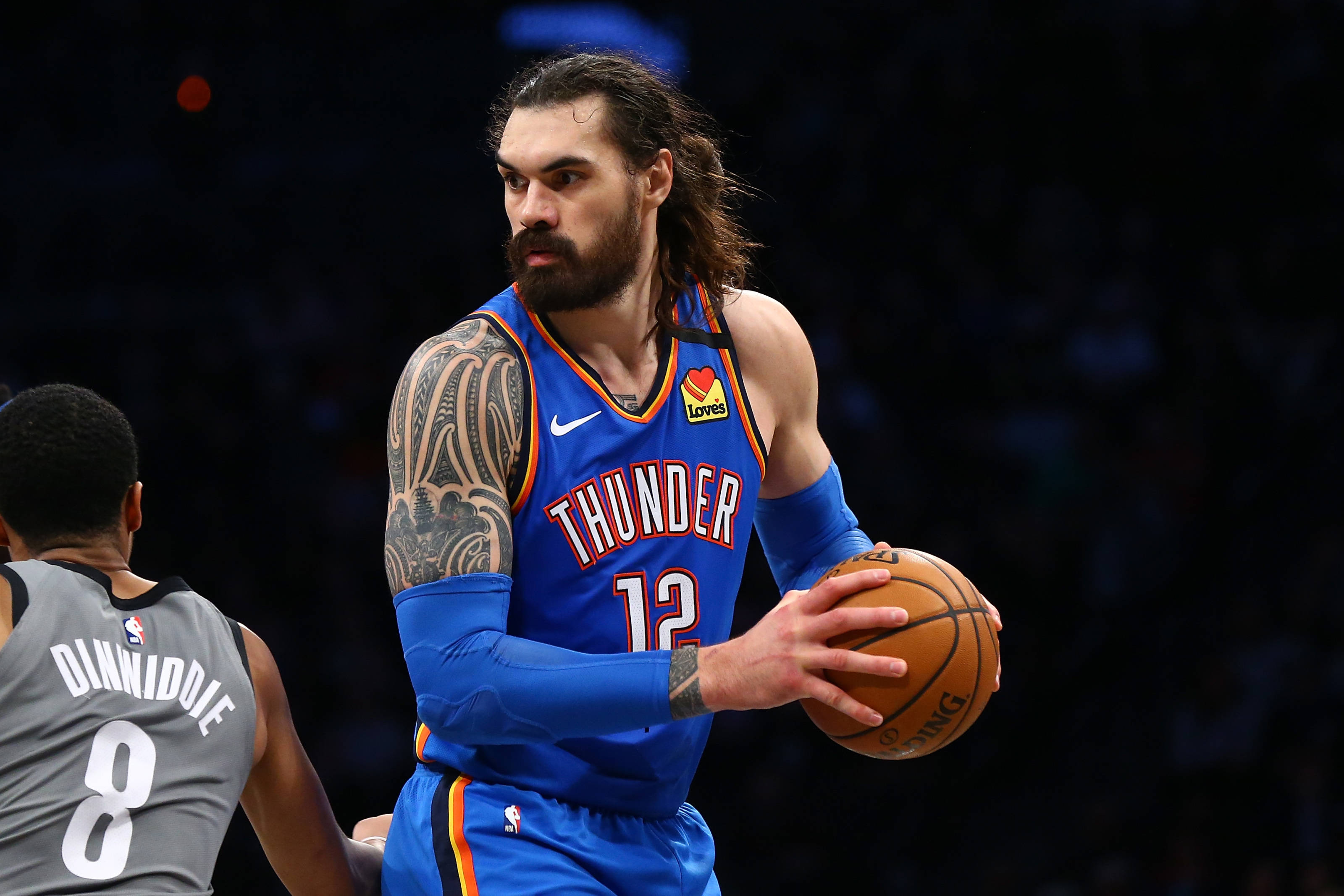 Steven Adams 桌布 壁紙 — 一位專注的籃球運動員身穿藍色雷霆球衣，在昏暗的球場上運球並被防守。