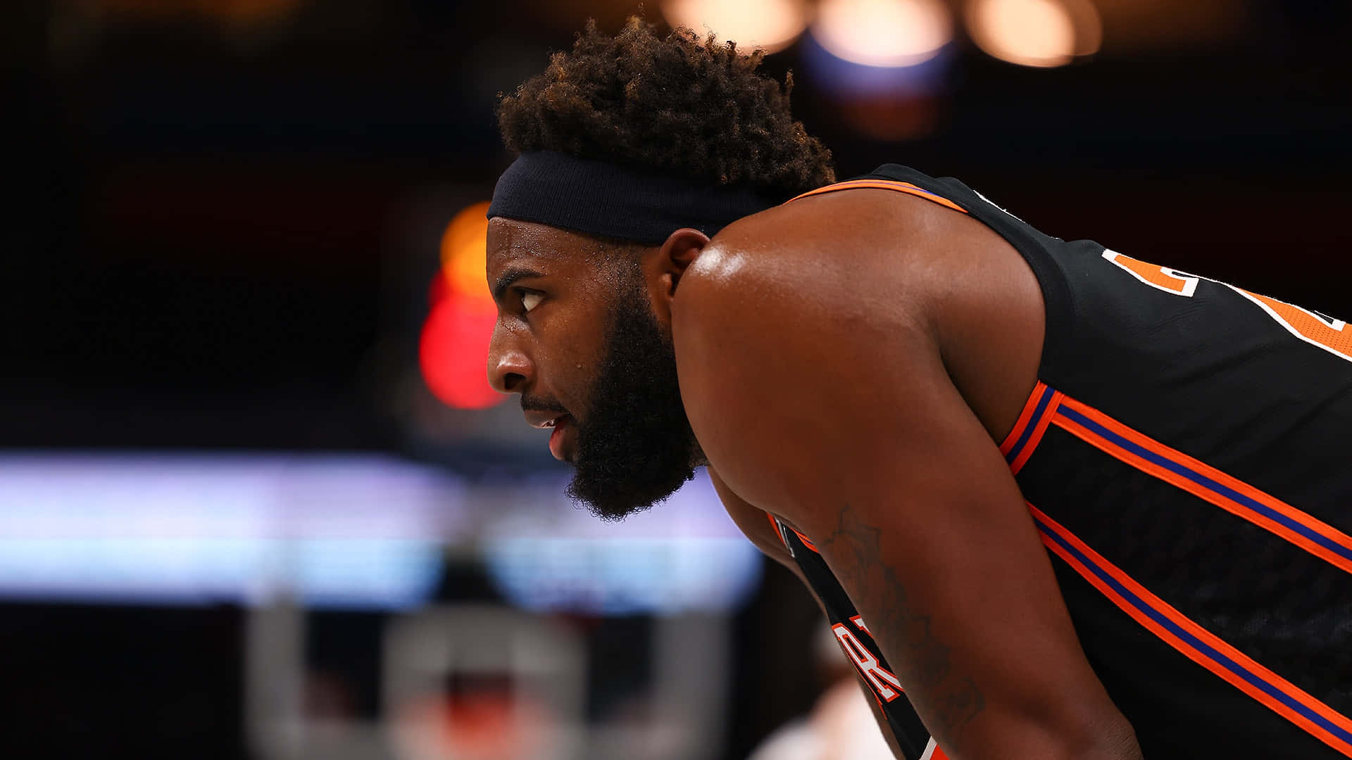 NBA 桌布 壁紙 — 一位專注的籃球運動員身穿黑色和橙色球衣，在明亮的場館燈光下比賽中激烈地向前傾。