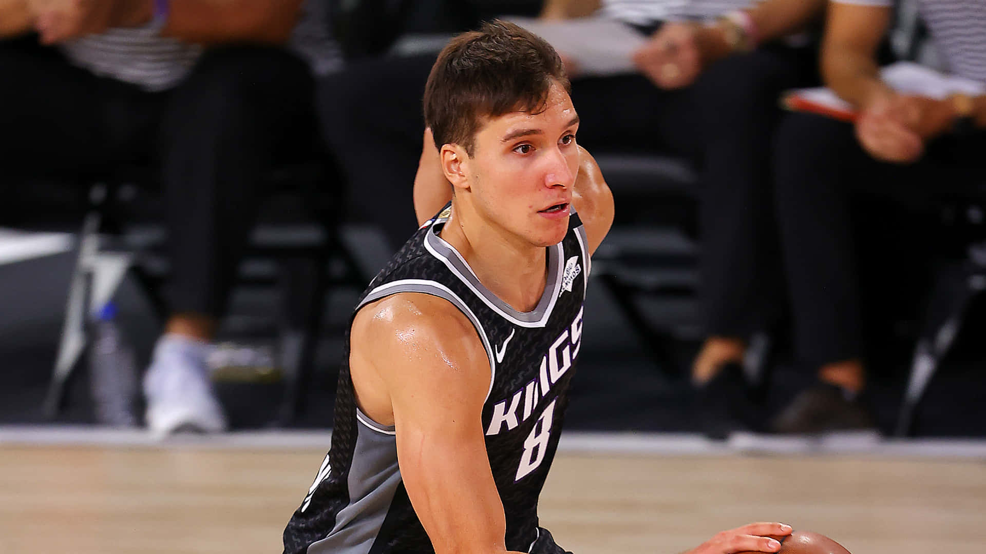 NBA 桌布 壁紙 — 一位專注的籃球運動員身穿黑色和銀色的Kings球衣，跪在拋光的地板上，背景觀眾模糊不清。
