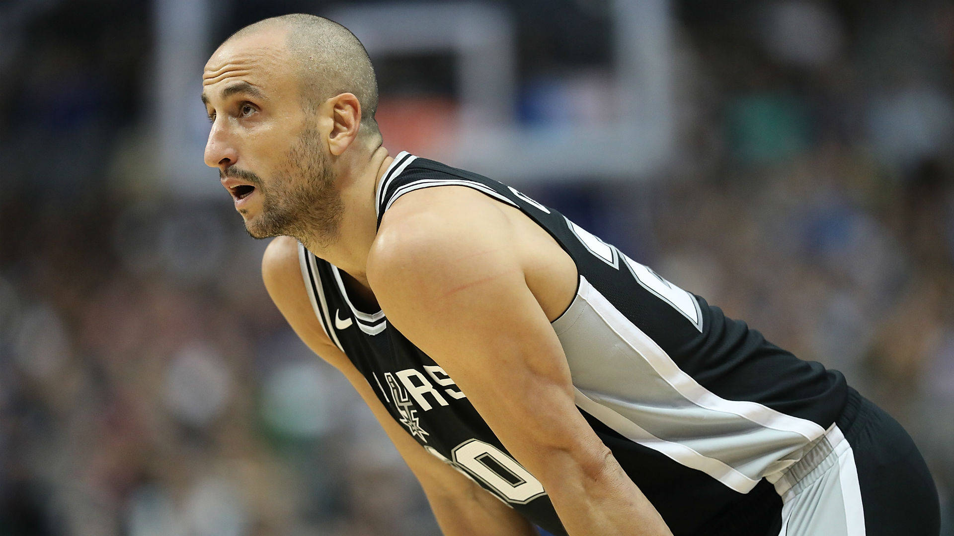 Manu 桌布 壁紙 — 一位專注的籃球運動員身穿黑色和銀色球衣，在明亮的場館燈光下於比賽中激烈地向前傾身。