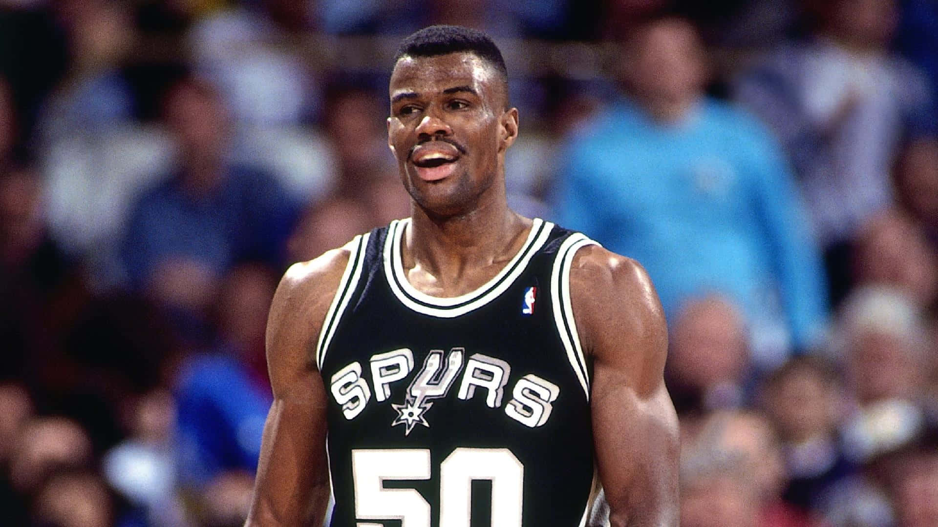 NBA 桌布 壁紙 — 一位專注的籃球運動員身穿黑色馬刺隊球衣，號碼50，站在球場上，背景是模糊的觀眾。