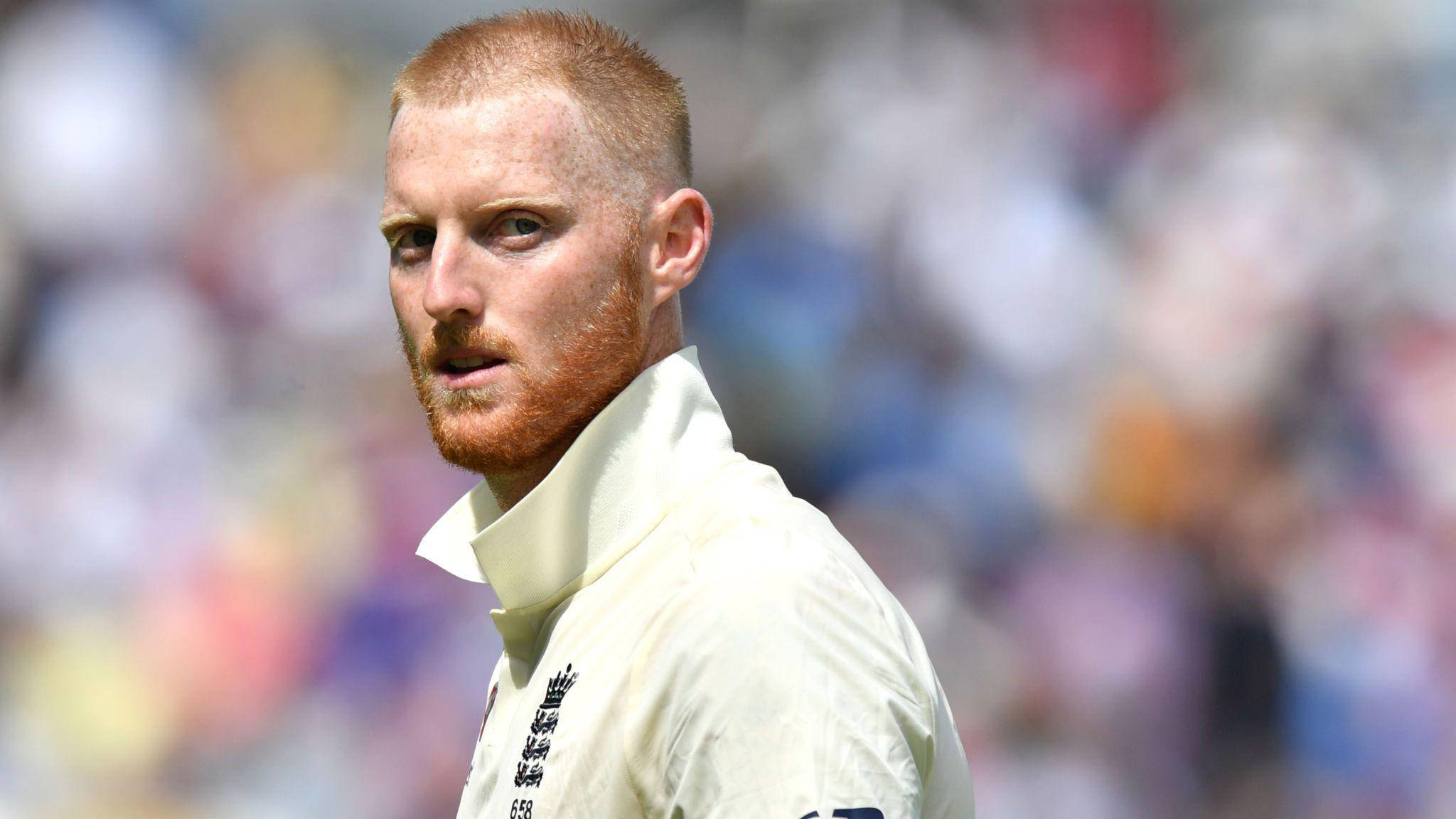Ben Stokes 桌布 壁紙 — 一位專注的紅髮板球運動員身穿白色制服，在明亮的日光下站在模糊的體育場人群前。