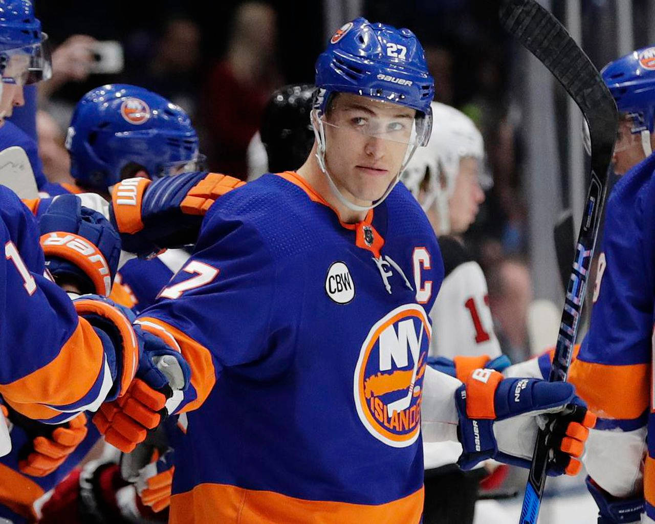 New York Islanders 桌布 壁紙 — 一位專注的紐約島人冰球球員，身穿藍色和橙色裝備，在比賽中手持球棍，隊友在附近。