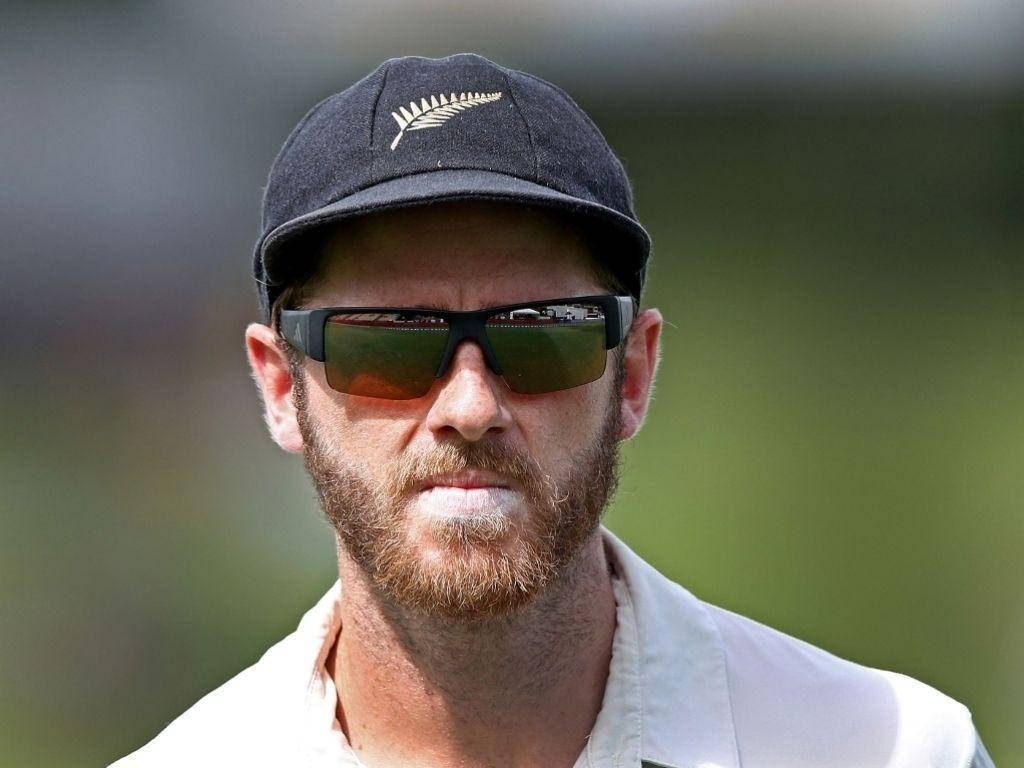 kane williamson 桌布 壁紙 — 一位專注的紐西蘭板球運動員，留著鬍鬚並戴著太陽眼鏡，在模糊的綠色場地上凝視。