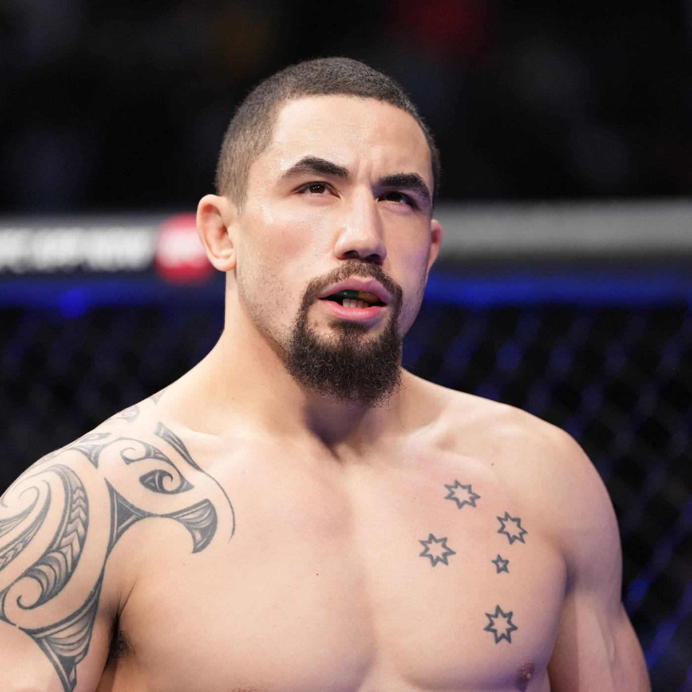 UFC 桌布 壁紙 — 一位專注的綜合格鬥家，身上有部落紋身和胸口星星紋身，站在燈光昏暗的八角籠內。
