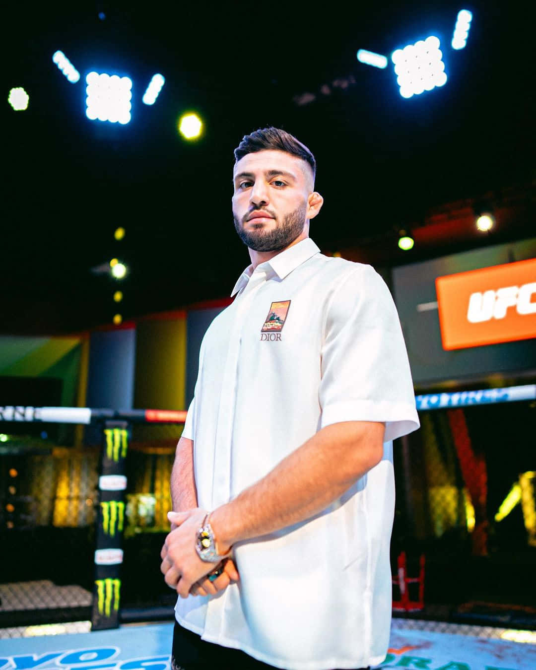 MMA 桌布 壁紙 — 一位專注的綜合格鬥家穿著白色Dior襯衫，自信地站在燈光明亮的UFC八角籠內。