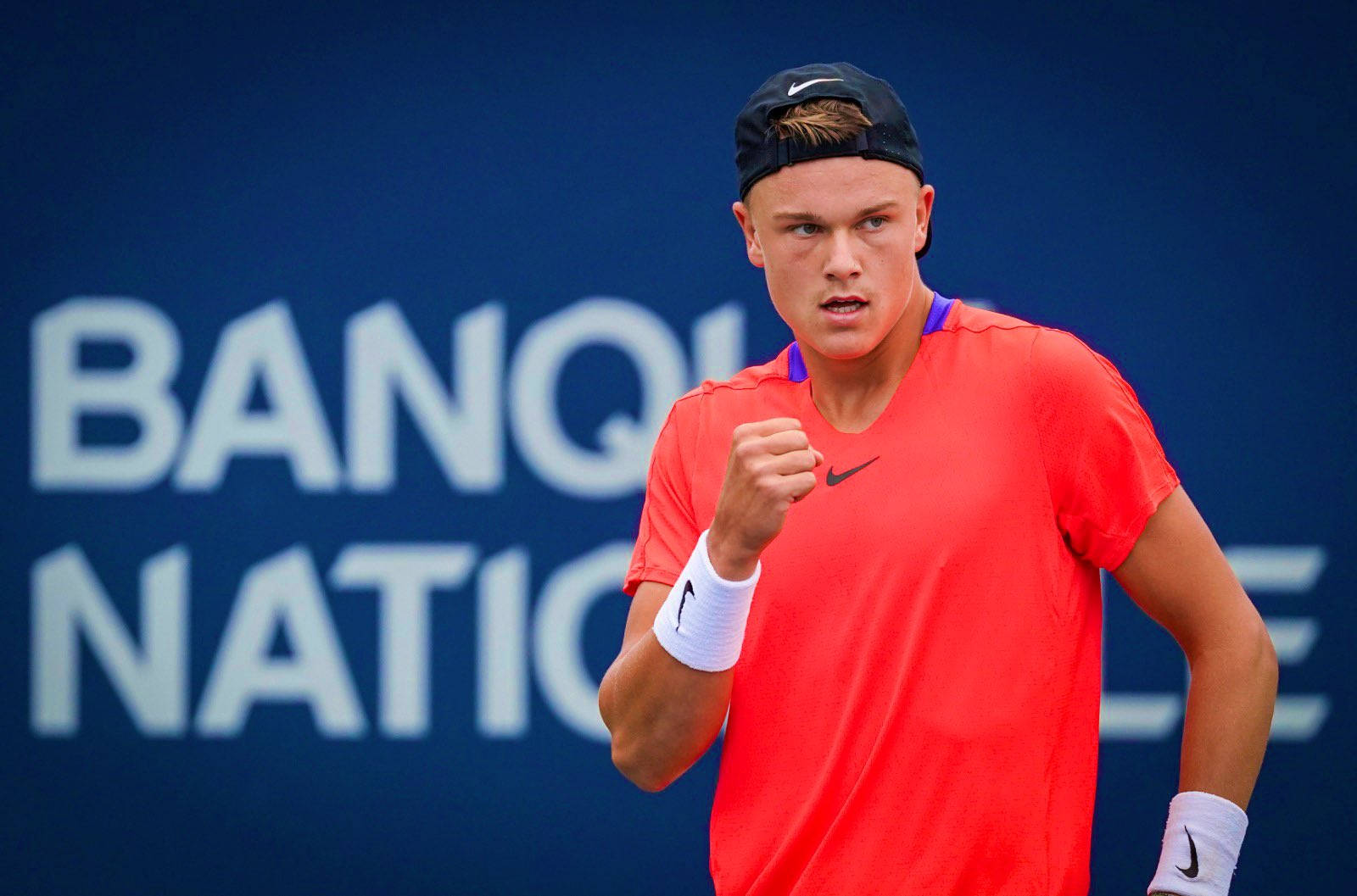 Tennis 桌布 壁紙 — 一位專注的網球運動員穿著亮橙色襯衫，在帶有藍色「BANQUE NATIONALE」背景的球場上握拳慶祝。