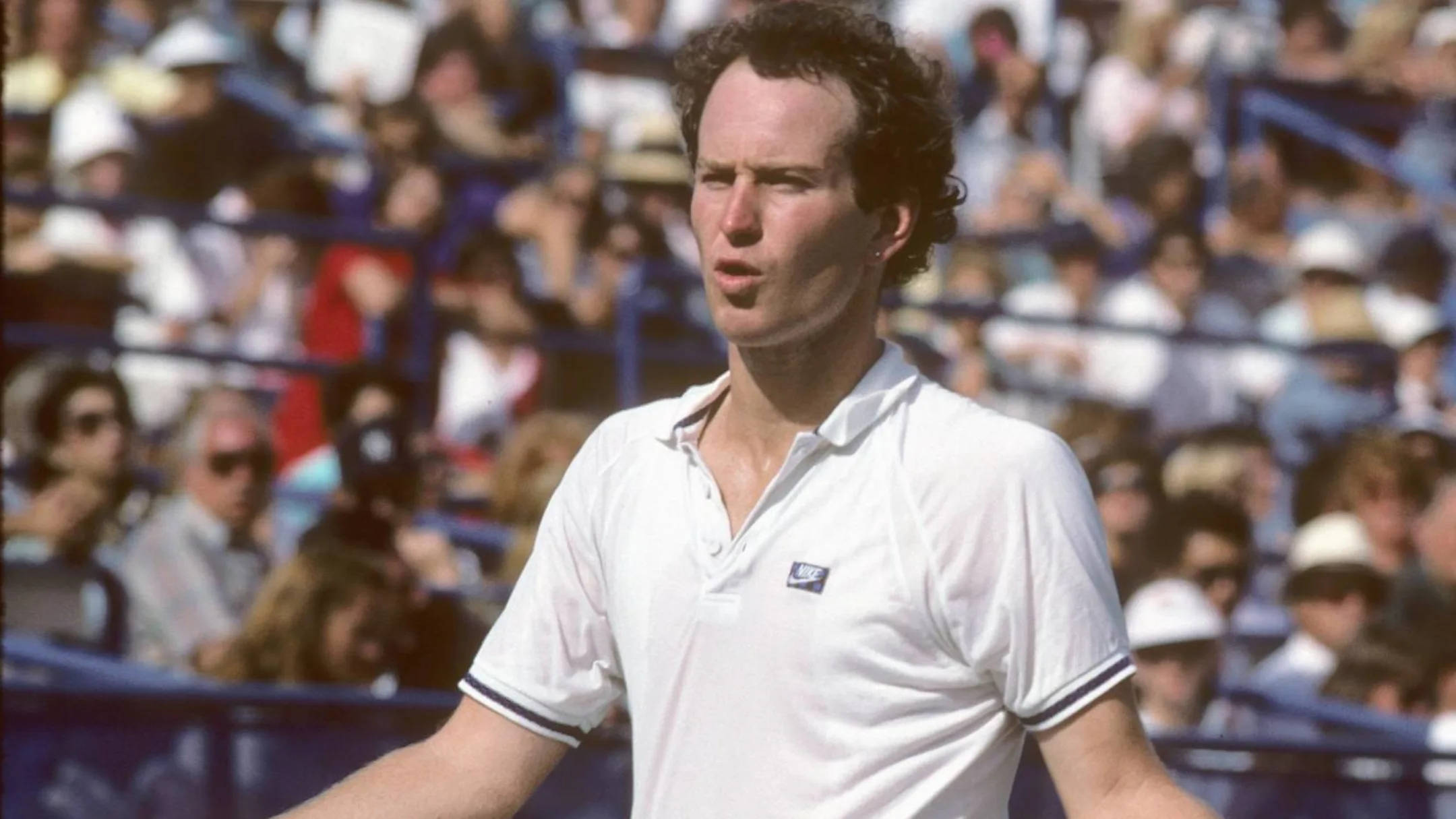 Tennis 桌布 壁紙 — 一位專注的網球運動員穿著白色Polo衫，站在球場上，背景有觀眾群。