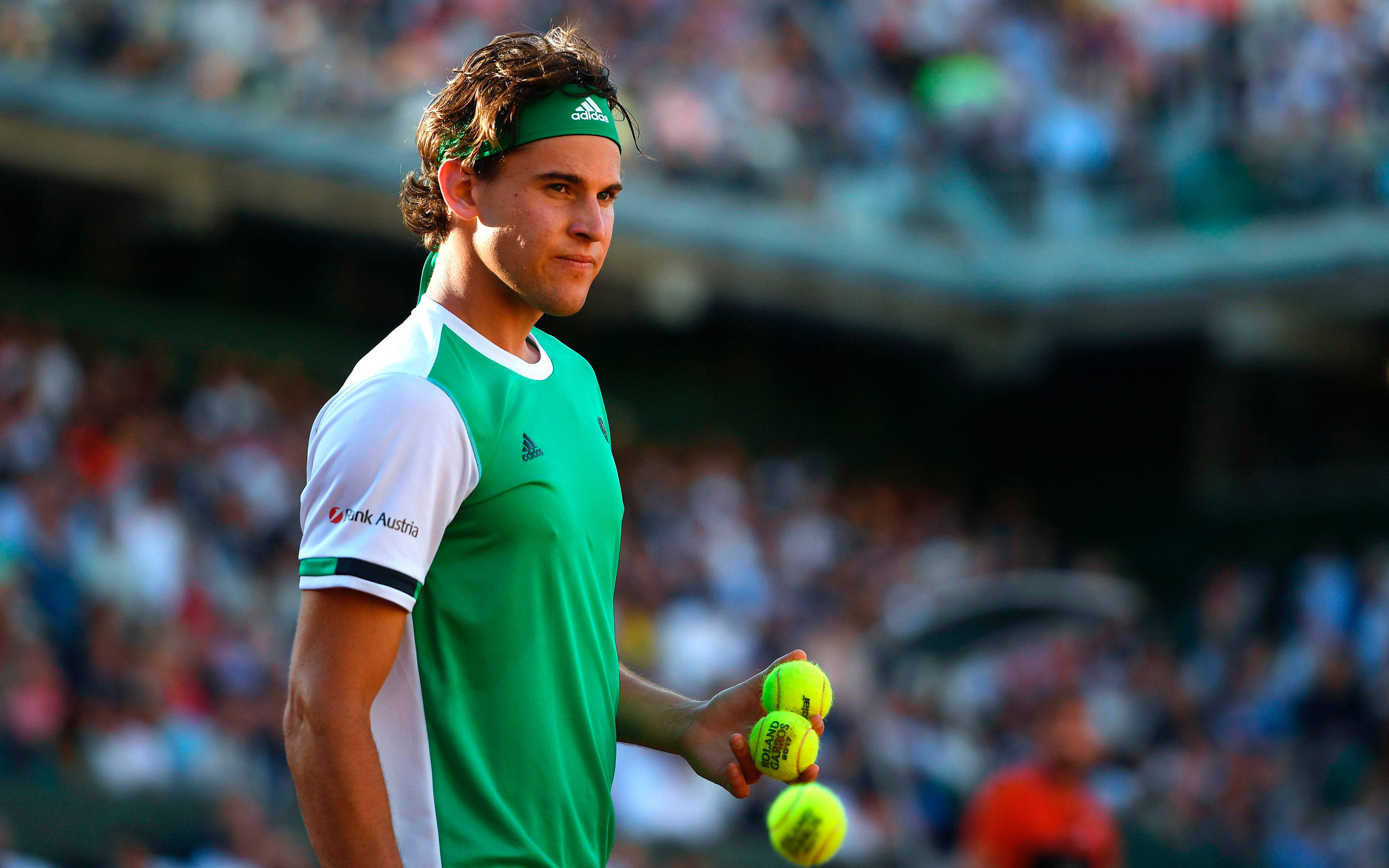 dominic thiem 桌布 壁紙 — 一位專注的網球選手身穿綠色和白色裝備，在陽光明媚的球場上拿著亮黃色的球，背景有模糊的觀眾。