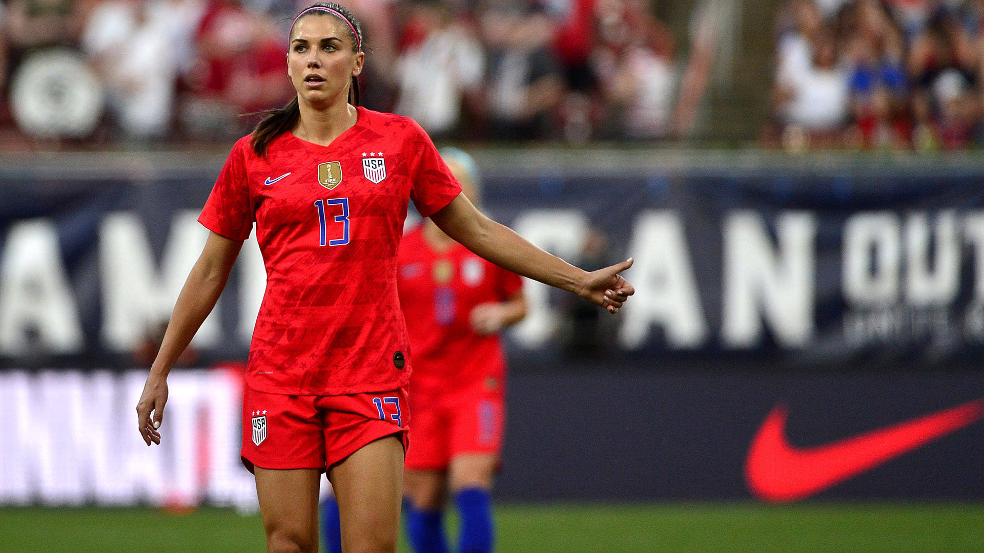 Alex Morgan 桌布 壁紙 — 一位專注的美國女足（USWNT）球員身穿紅色13號球衣，在比賽期間於充滿活力的綠色場地上做手勢。