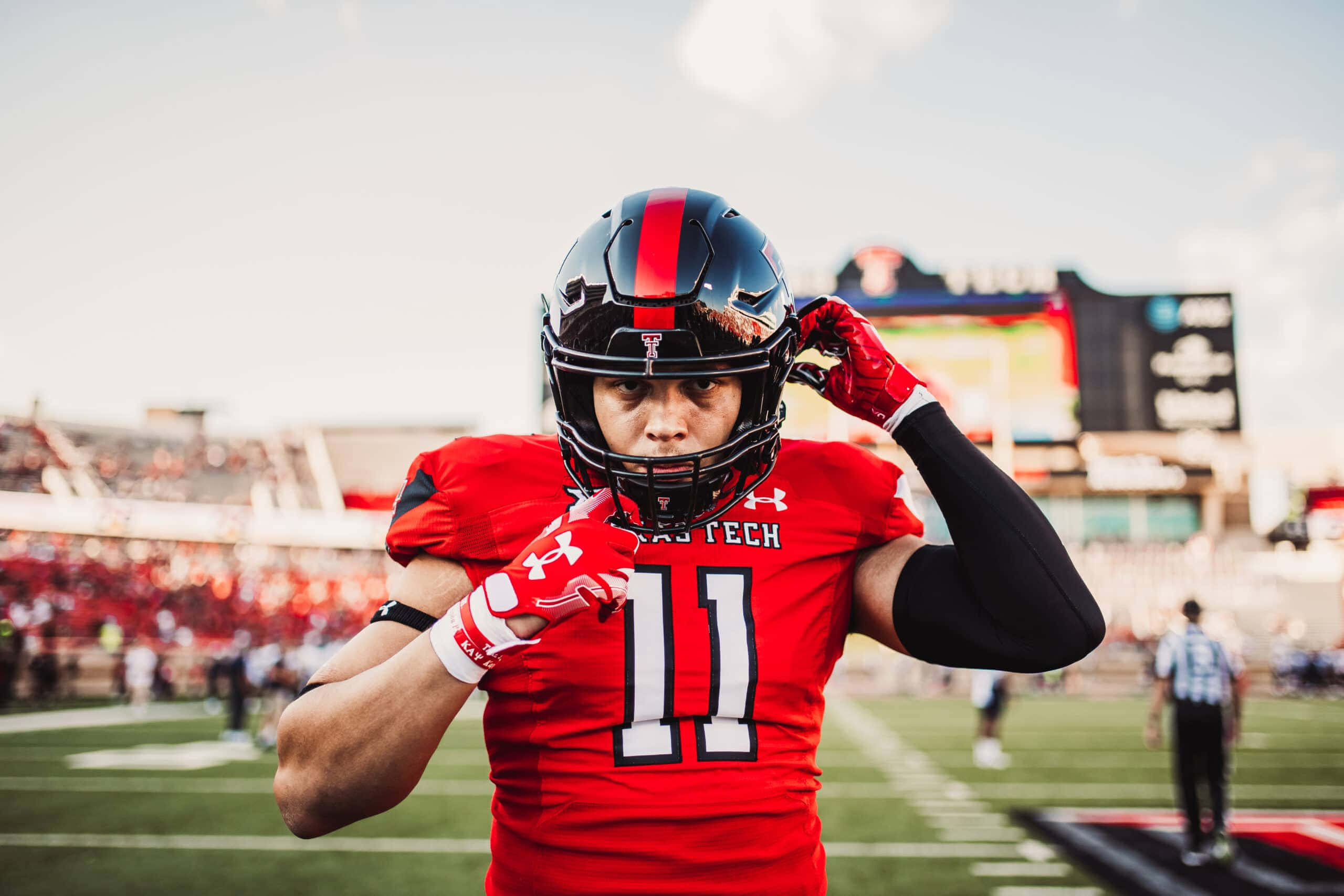 Texas Tech 桌布 壁紙 — 一位專注的美式足球員穿著紅色德州理工大學球衣，在陽光明媚的球場上調整頭盔，背景是體育場的人群。