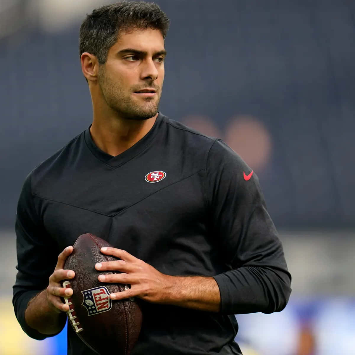 Jimmy Garoppolo 桌布 壁紙 — 一位專注的美式足球員身穿黑色舊金山49人隊球衣，在模糊的球場背景下拿著一個棕色的NFL足球。