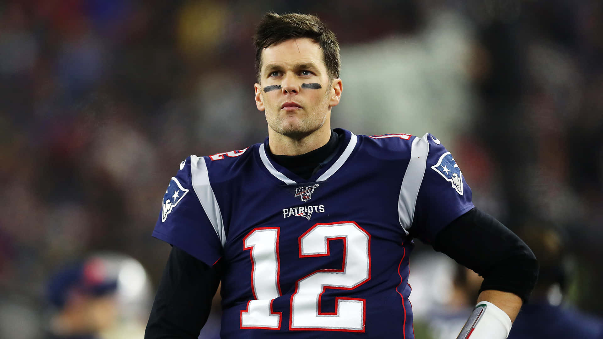 New England Patriots 桌布 壁紙 — 一位專注的美式足球四分衛，身穿新英格蘭愛國者隊球衣，帶著堅定的表情站在球場上。