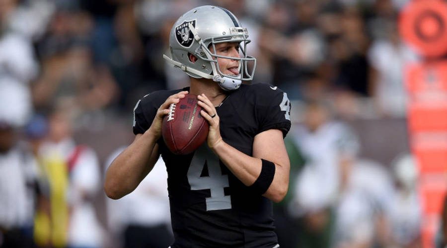Football Players 桌布 壁紙 — 一位專注的美式足球四分衛，身穿黑色球衣和銀色頭盔，在日間比賽中準備投球。