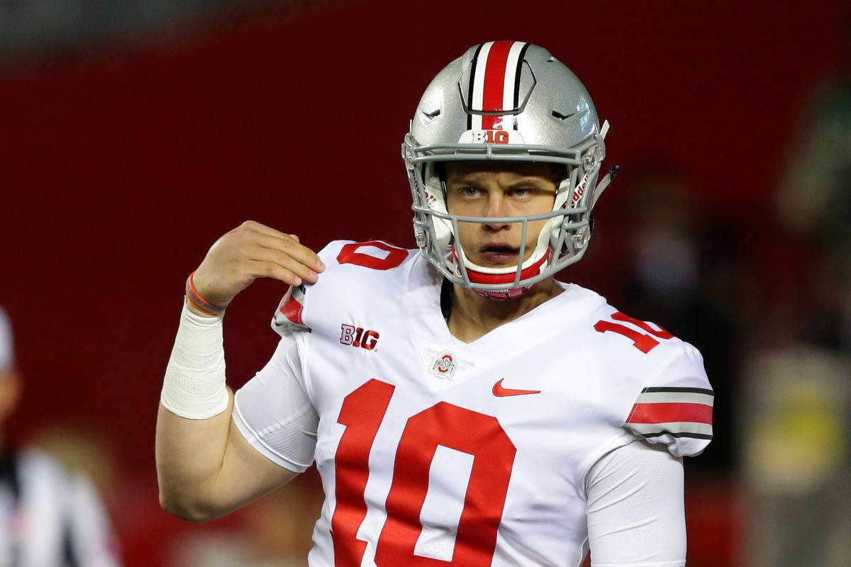 Joe Burrow 桌布 壁紙 — 一位專注的美式足球四分衛穿著白色和紅色制服，在體育場燈光下調整頭盔。