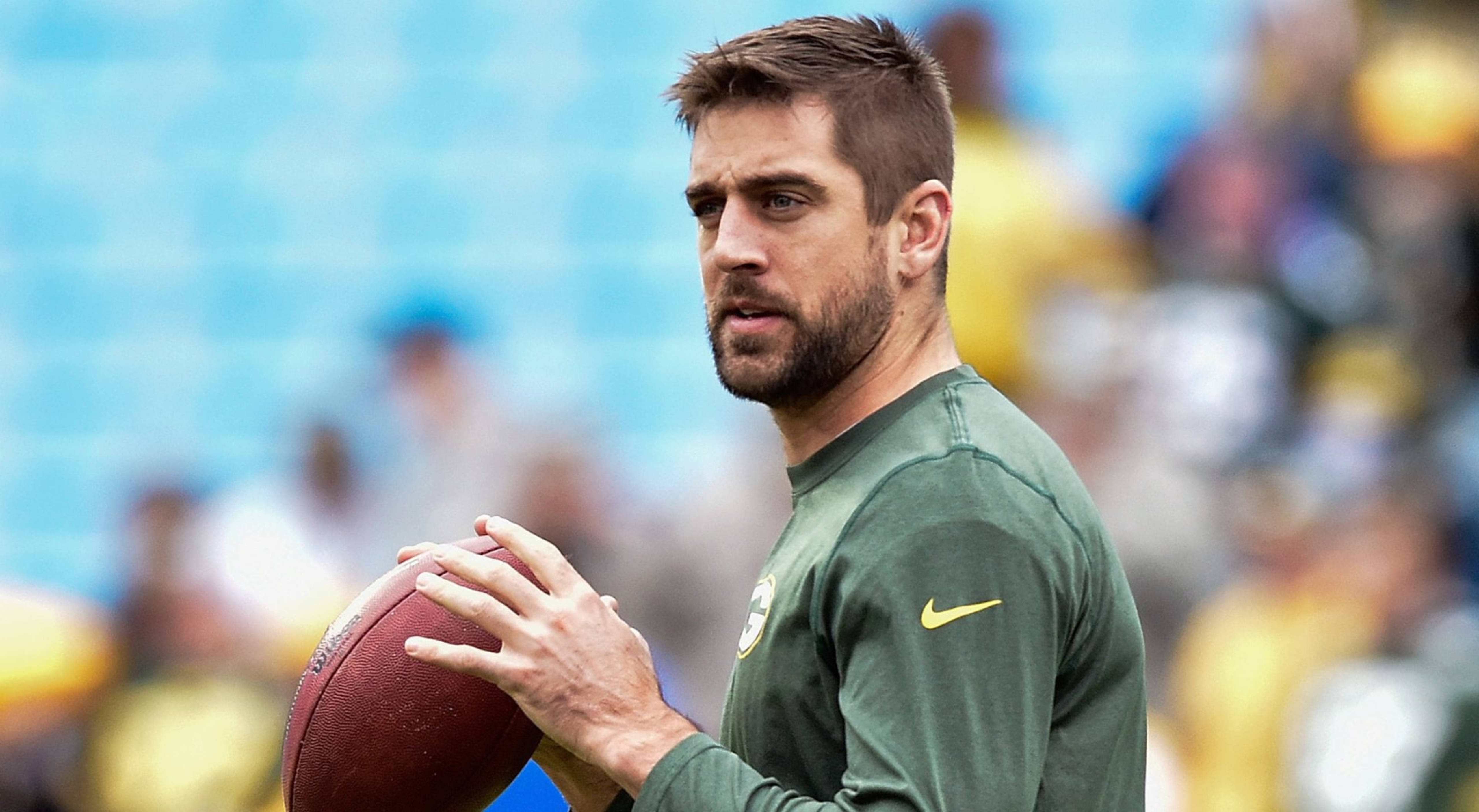 Aaron Rodgers 桌布 壁紙 — 一位專注的美式足球四分衛穿著綠色球衣，拿著棕色美式足球，站在球場上，身後是模糊的體育場座位。