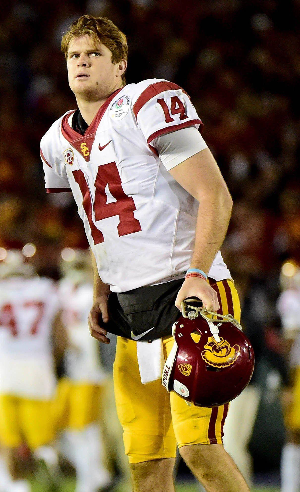 Football Players 桌布 壁紙 — 一位專注的美式足球四分衛身穿白色和金色制服，在夜間比賽中於體育場燈光下拿著頭盔向上看。