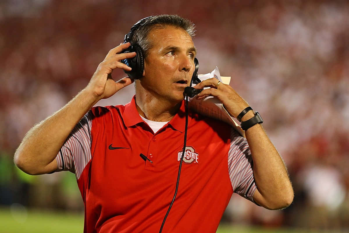 Urban Meyer 桌布 壁紙 — 一位專注的美式足球教練，身穿紅色俄亥俄州立大學Polo衫，在夜間燈光球場上調整耳機。