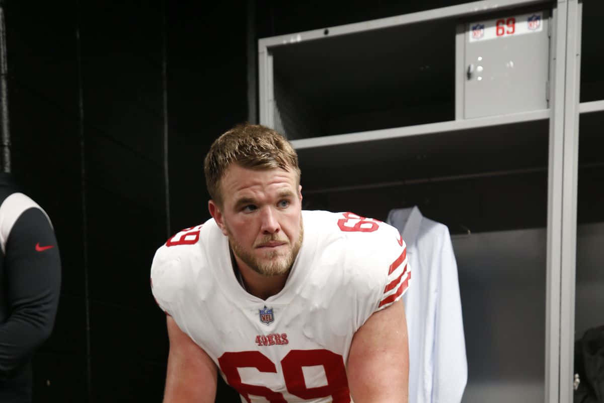 Mike Mcglinchey 桌布 壁紙 — 一位專注的舊金山49人美式足球員，身穿白色69號球衣，在昏暗的更衣室裡向前傾。