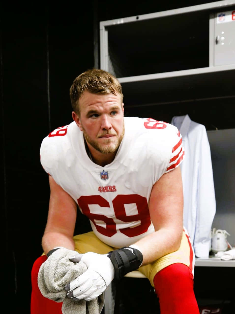 Mike Mcglinchey 桌布 壁紙 — 一位專注的舊金山49人美式足球員，身穿69號球衣，坐在燈光昏暗的更衣室裡，拿著毛巾。