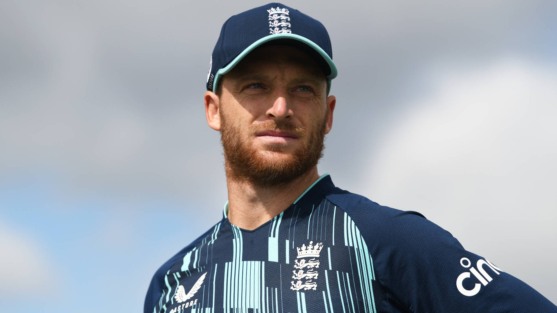 jos buttler 桌布 壁紙 — 一位專注的英格蘭板球運動員穿著隊服，在陰雲密布的天空下帶著堅定的目光站立。