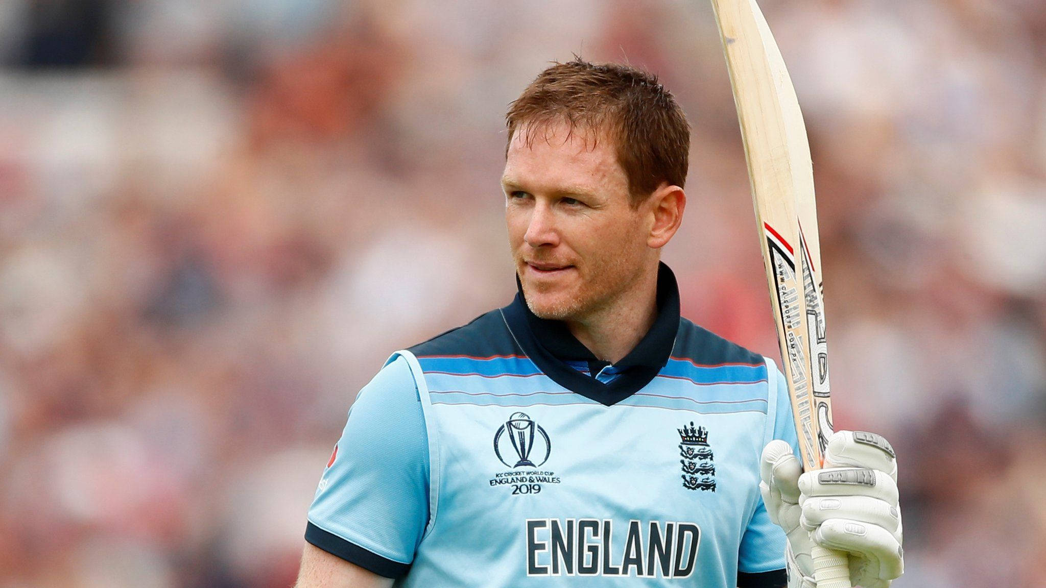 eoin morgan 桌布 壁紙 — 一位專注的英格蘭板球運動員身穿淺藍色球衣，手持球棒，在陽光明媚的球場上行走，身後有模糊的觀眾。