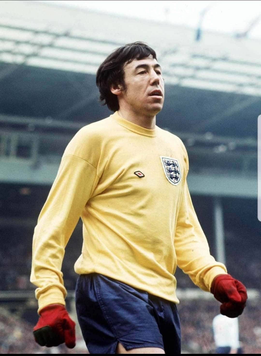 Athlete 桌布 壁紙 — 一位專注的英格蘭足球守門員，身穿復古黃色球衣和紅色手套，警覺地站在球場上。