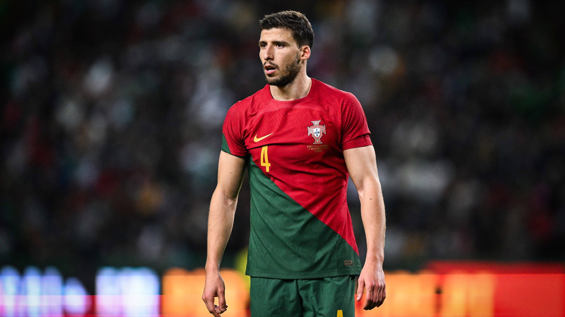 Football Players 桌布 壁紙 — 一位專注的葡萄牙足球員身穿紅綠球衣，在比賽期間站在燈光明亮的球場上。