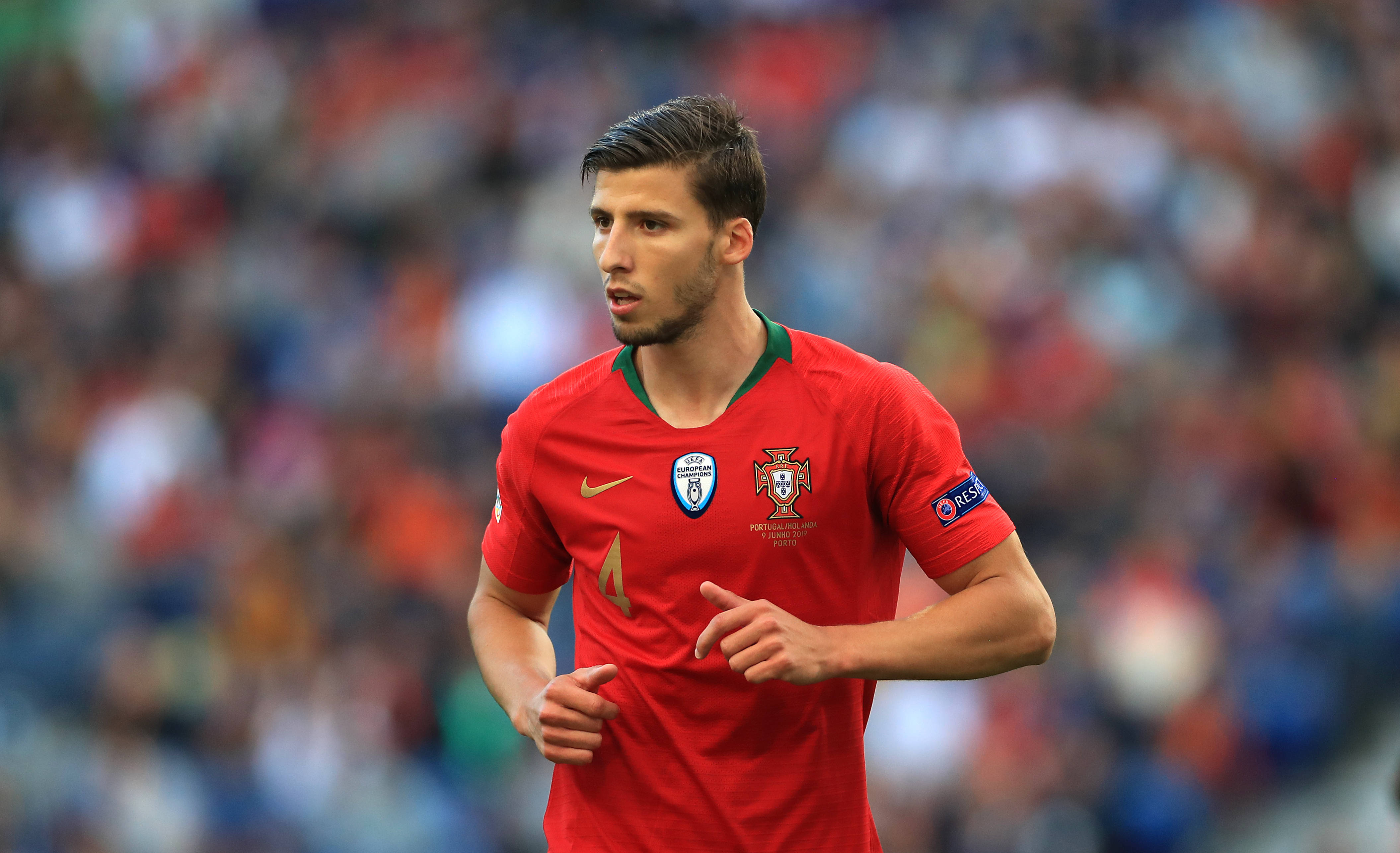 Football Players 桌布 壁紙 — 一位專注的葡萄牙足球員身穿紅色球衣，在日間比賽中奔跑，背景是模糊的體育場人群。