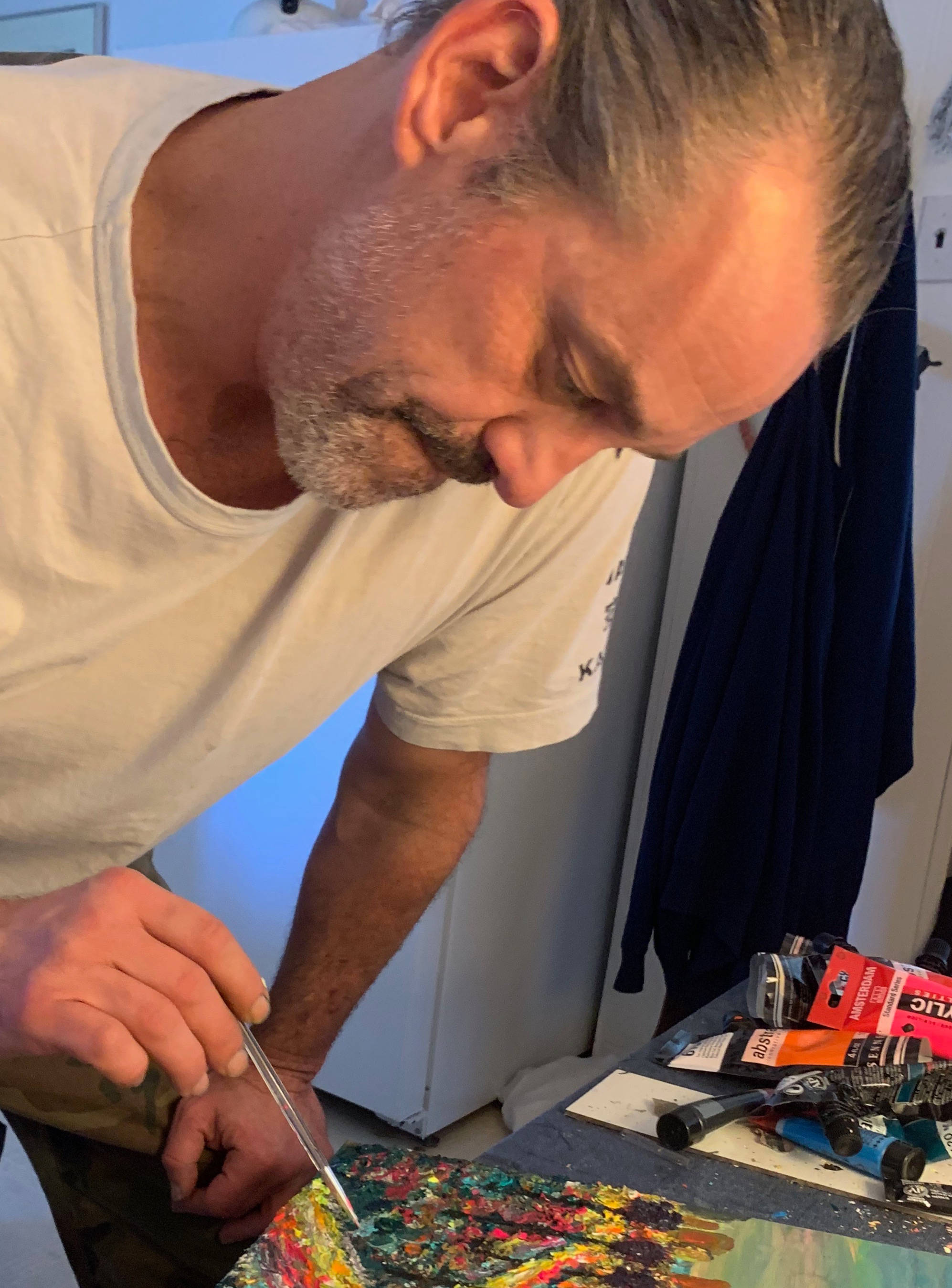Nicholas Brendon 桌布 壁紙 — 一位專注的藝術家，頭髮花白，在雜亂的工作室裡用調色刀在充滿活力的抽象畫上彎腰作畫。