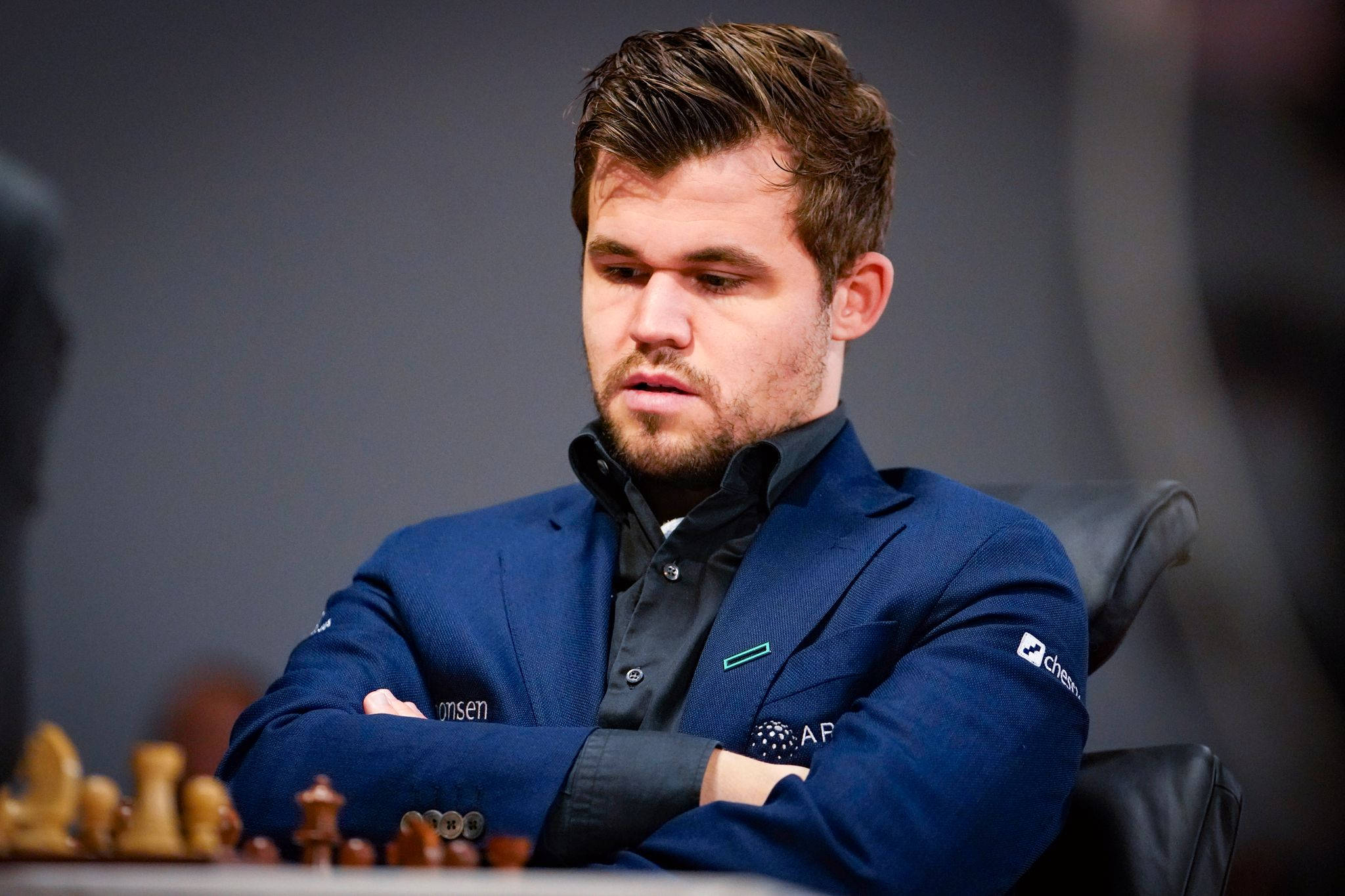 Magnus 桌布 壁紙 — 一位專注的西洋棋棋手身穿藍色西裝外套，在昏暗的房間裡雙臂交叉俯身在棋盤上。
