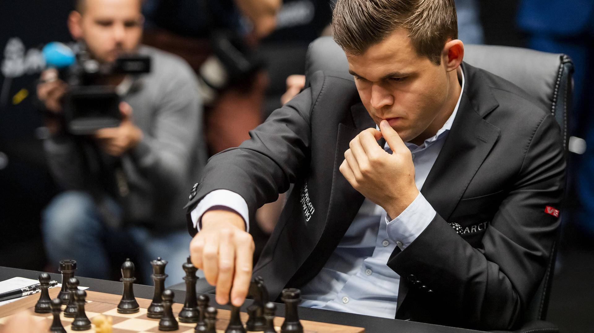 Magnus 桌布 壁紙 — 一位專注的西洋棋選手穿著黑色西裝，在木製棋盤上移動棋子，攝影師捕捉這緊張的時刻。