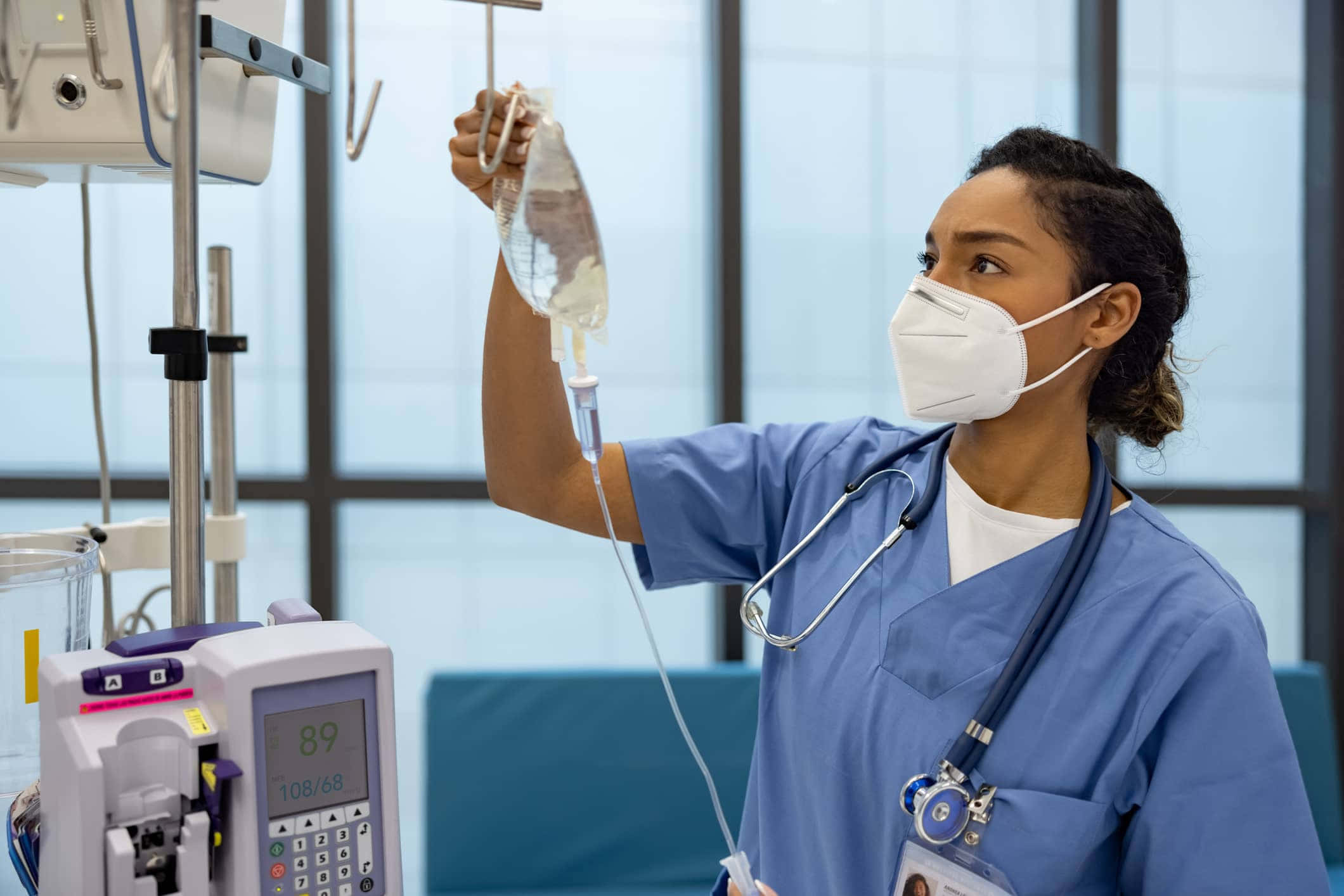 Nursing 桌布 壁紙 — 一位專注的護士穿著藍色工作服，在明亮的病房裡調整醫療設備旁的靜脈輸液袋。