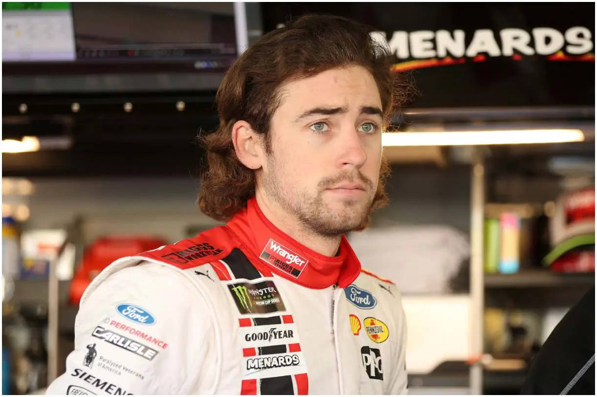 Ryan Blaney 桌布 壁紙 — 一位專注的賽車手穿著白色、紅色和黑色贊助商覆蓋的制服，站在車庫中，身後有模糊的設備和標誌。