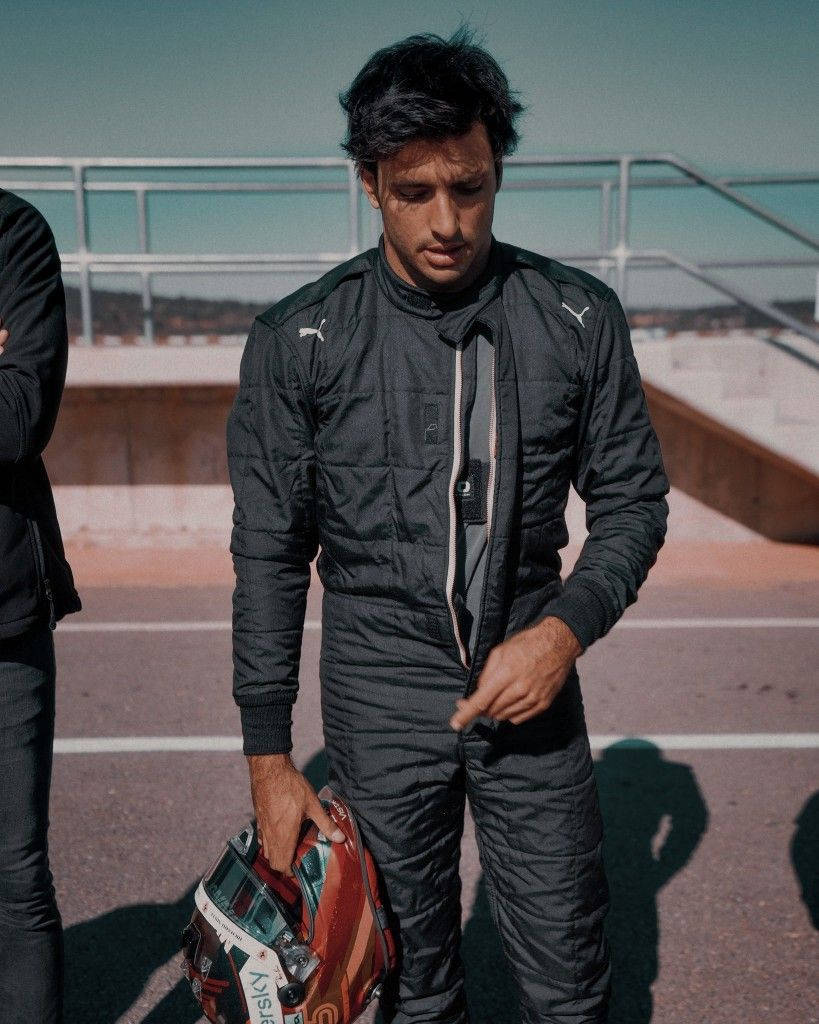 Racing 桌布 壁紙 — 一位專注的賽車手身穿黑色賽車服，站在陽光普照的賽道旁平台上，手持頭盔。