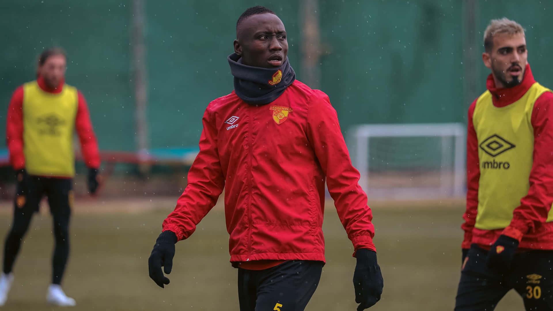 Football Players 桌布 壁紙 — 一位專注的足球員穿著紅色訓練外套，在潮濕的場地上與穿著黃色訓練背心的隊友一起進行雨中訓練。