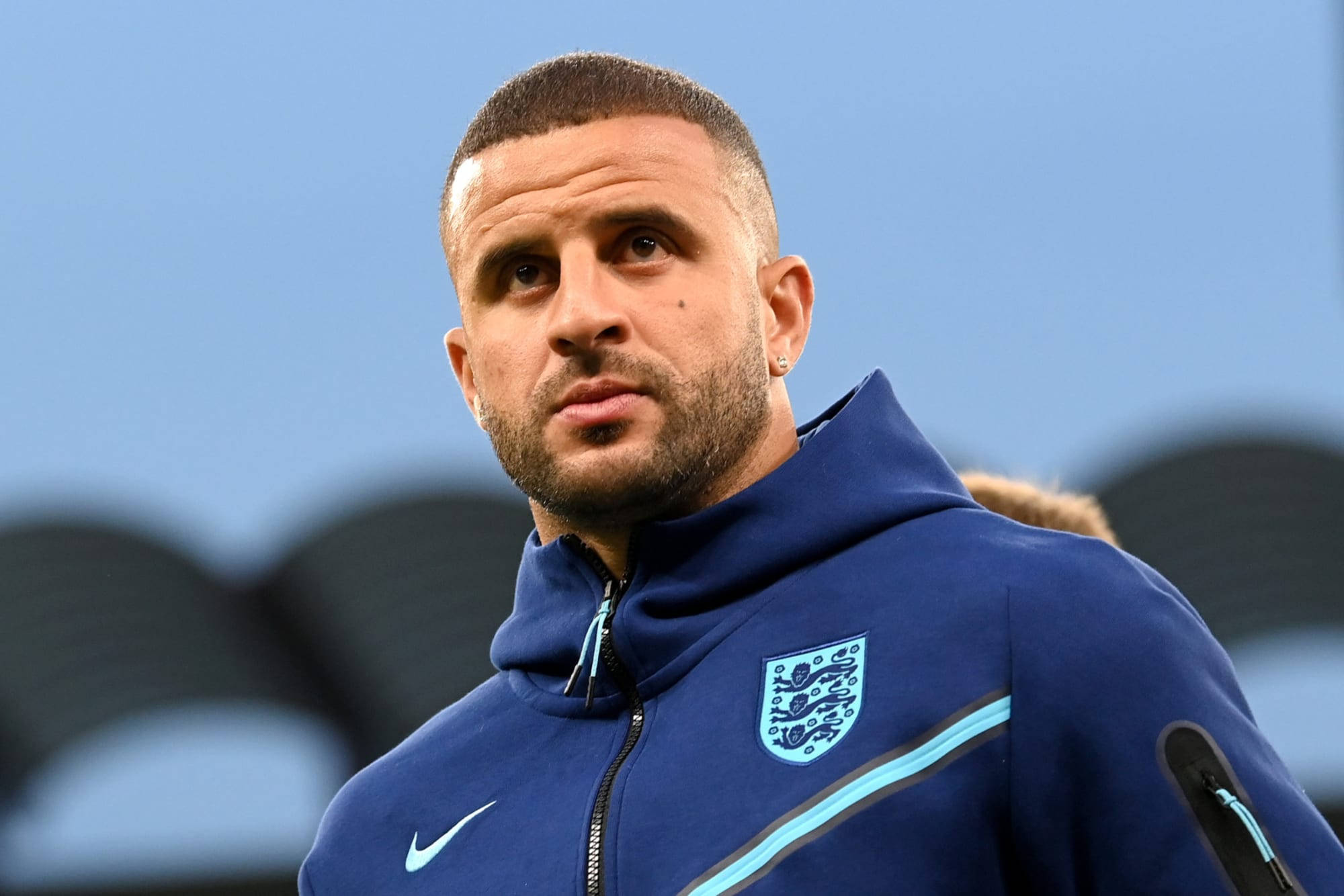 Kyle Walker 桌布 壁紙 — 一位專注的足球員穿著藍色英格蘭訓練外套，在模糊的體育場背景下凝視著。