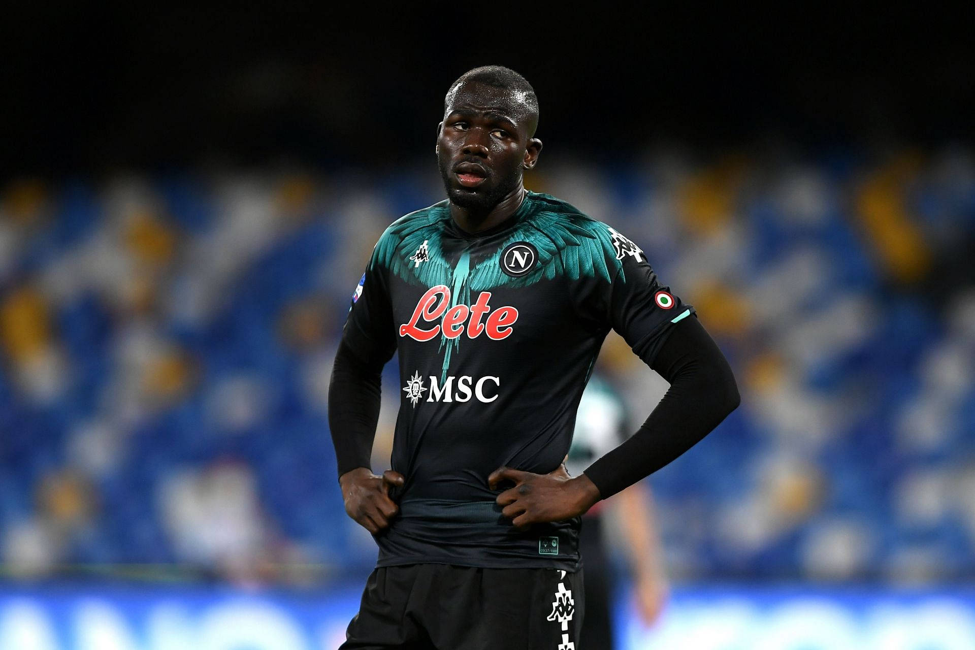 Athletic 桌布 壁紙 — 一位專注的足球員身穿深藍綠色和黑色的拿坡里球衣，在明亮的燈光下雙手叉腰站在球場上。