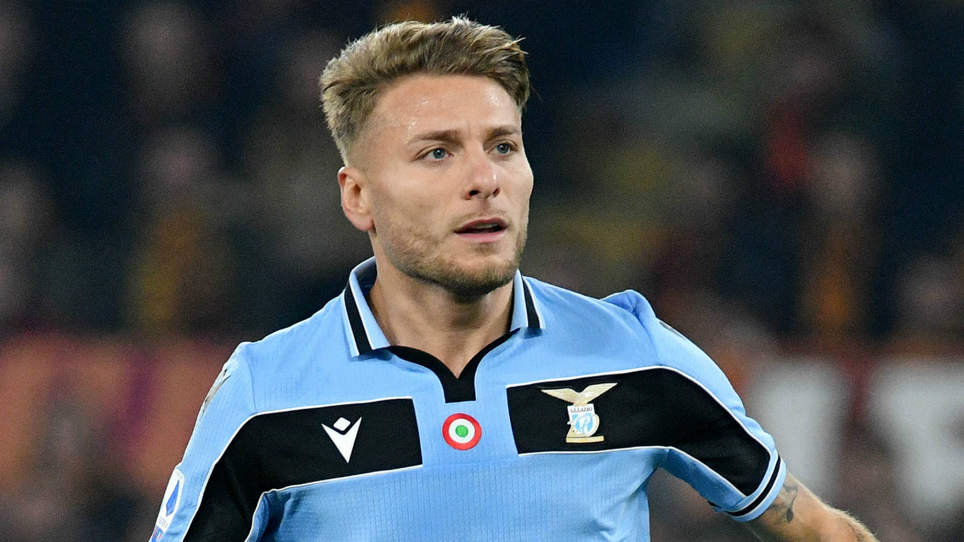 ciro immobile 桌布 壁紙 — 一位專注的足球員身穿淺藍色和黑色球衣，在球場燈光下比賽時專注地向前看。