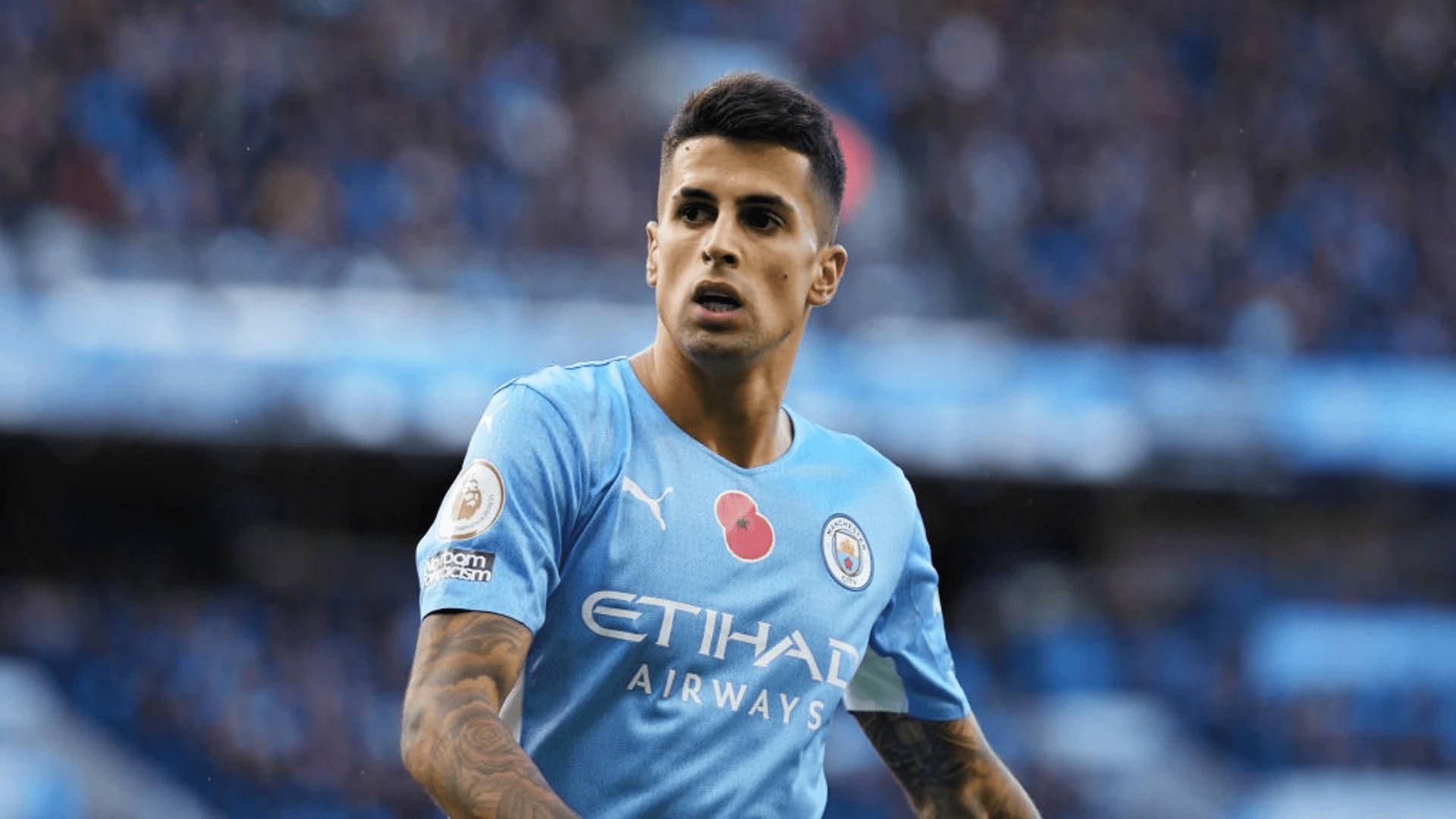 Football Players 桌布 壁紙 — 一位專注的足球員身穿淺藍色曼城球衣，手臂上有刺青，站在球場上。