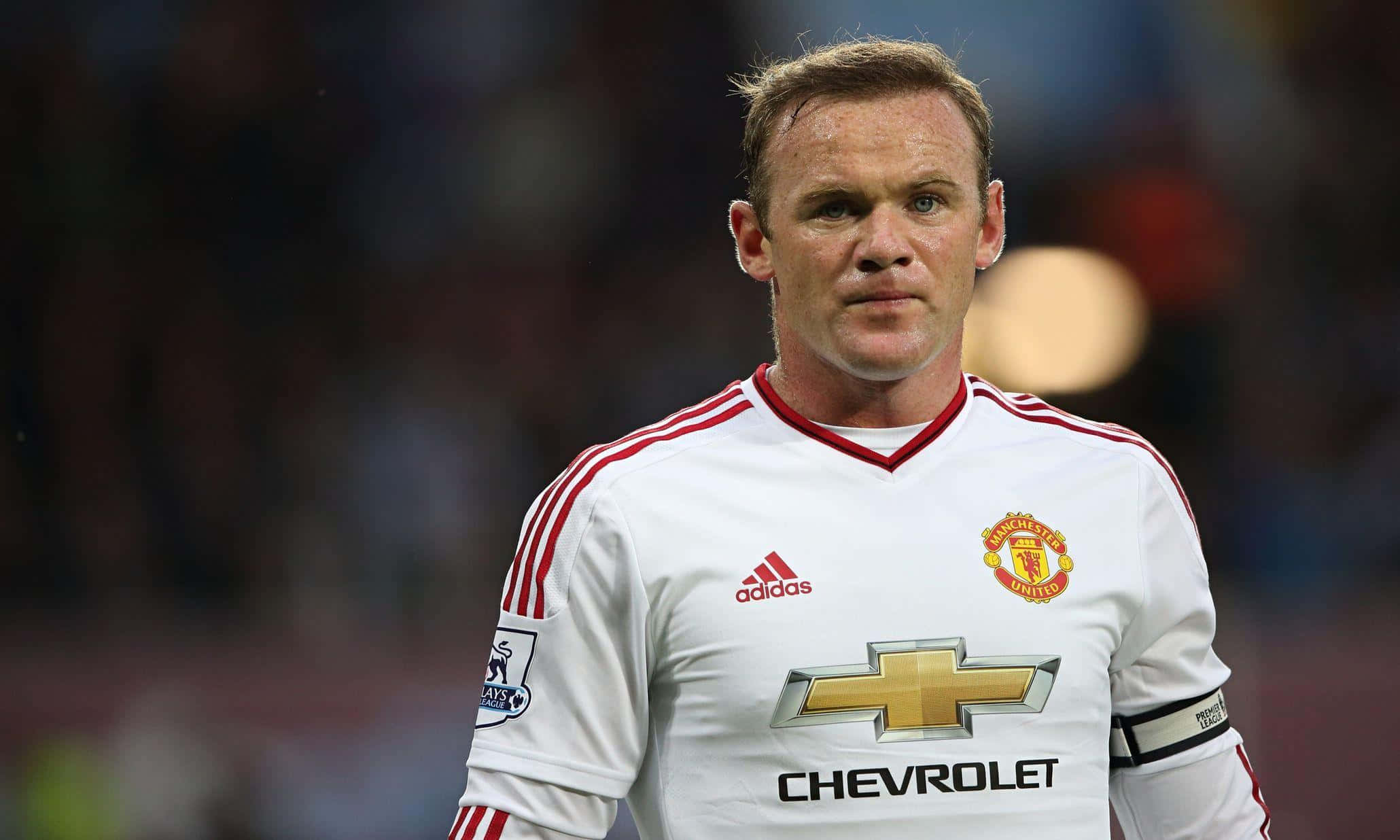 Rooney 桌布 壁紙 — 一位專注的足球員身穿白色曼聯球衣，帶有紅色條紋，站在燈光明亮的模糊球場上。