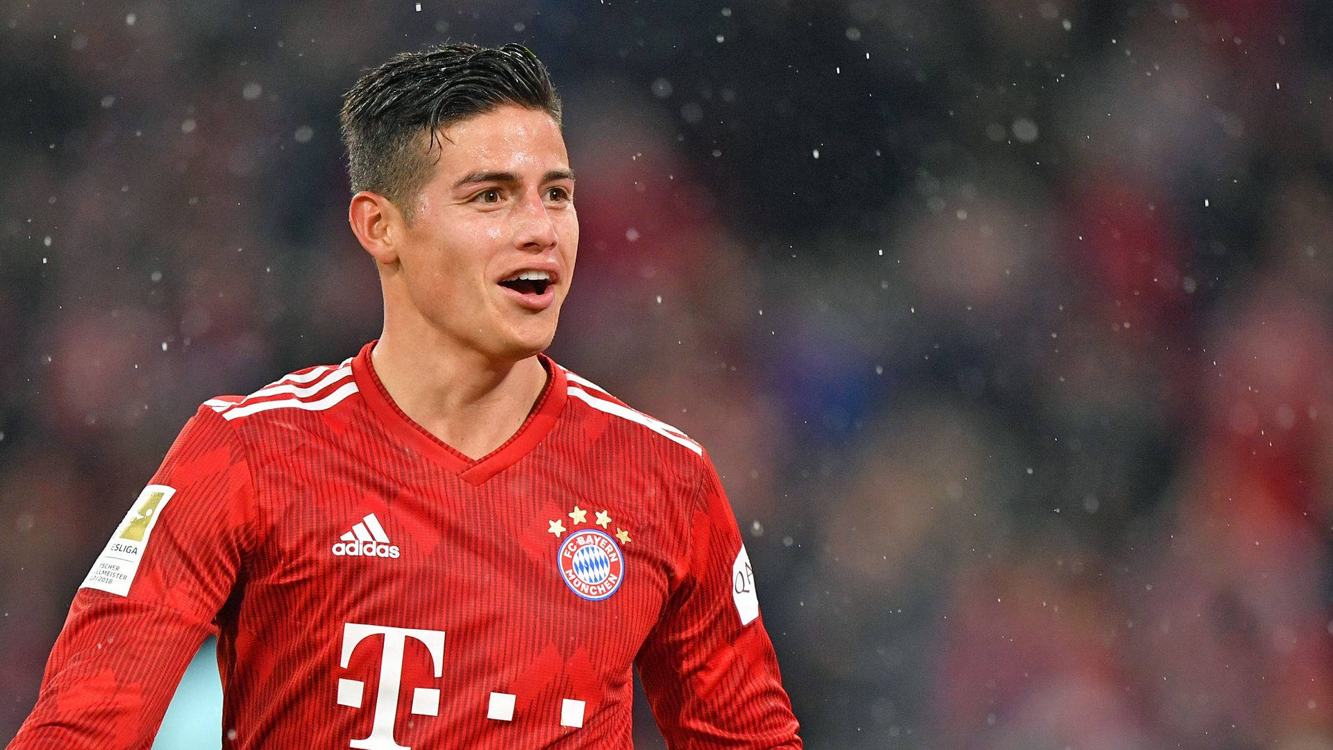 James Rodriguez 桌布 壁紙 — 一位專注的足球員身穿紅色拜仁慕尼黑球衣，站在雨中的球場上，身後是模糊的球場燈光。