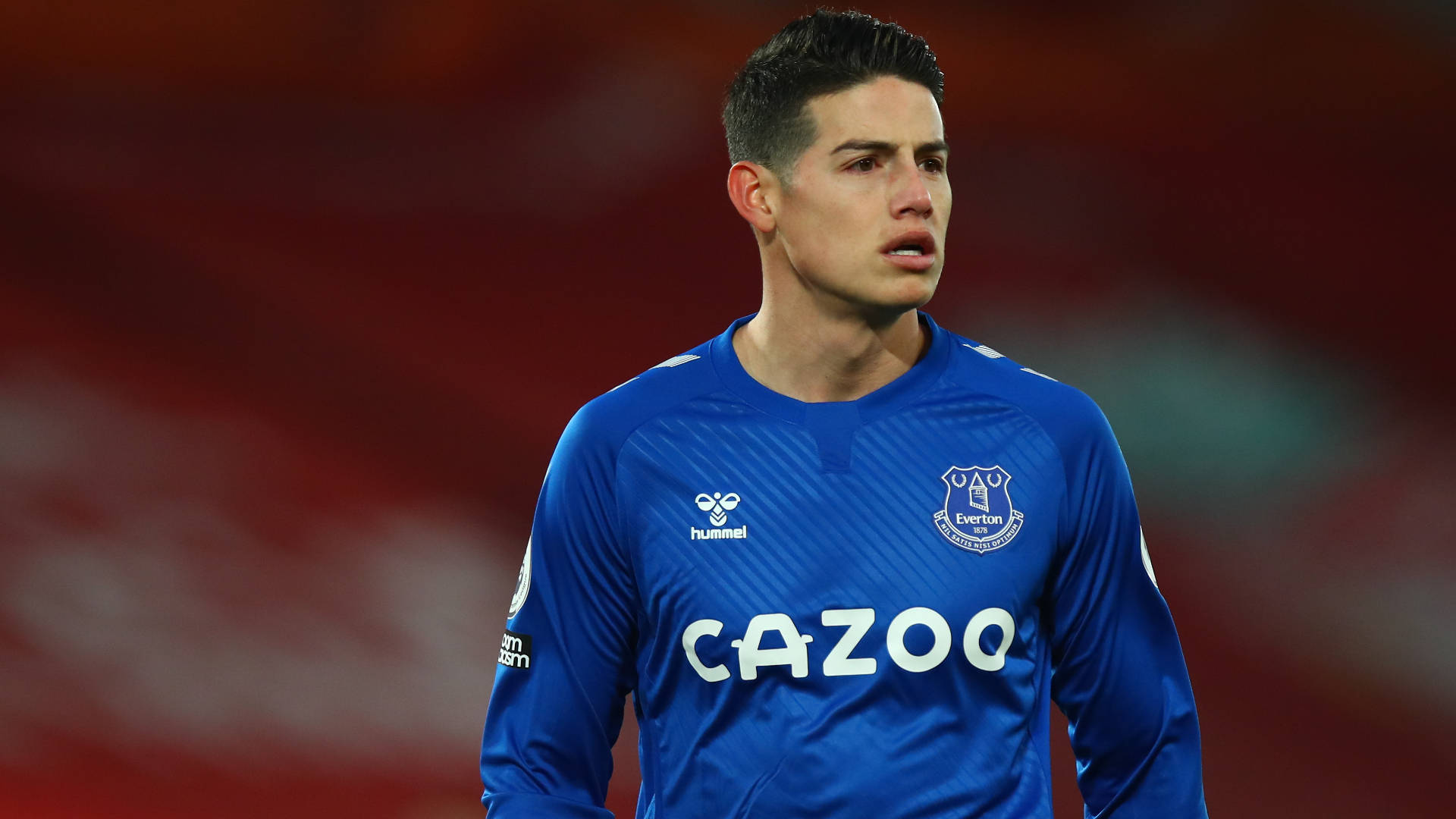 James Rodriguez 桌布 壁紙 — 一位專注的足球員身穿藍色艾佛頓球衣，站在模糊的紅色球場背景前。