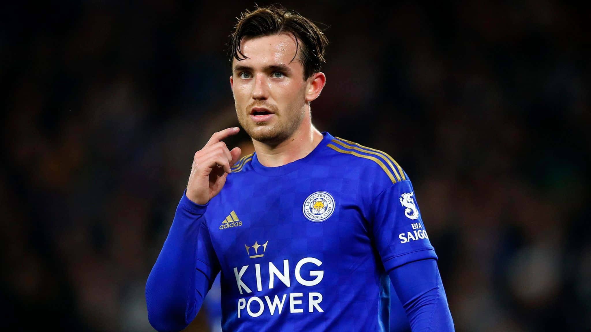 Football Players 桌布 壁紙 — 一位專注的足球員身穿藍色萊斯特城球衣，上面有「King Power」字樣，在夜間比賽中若有所思地做手勢。