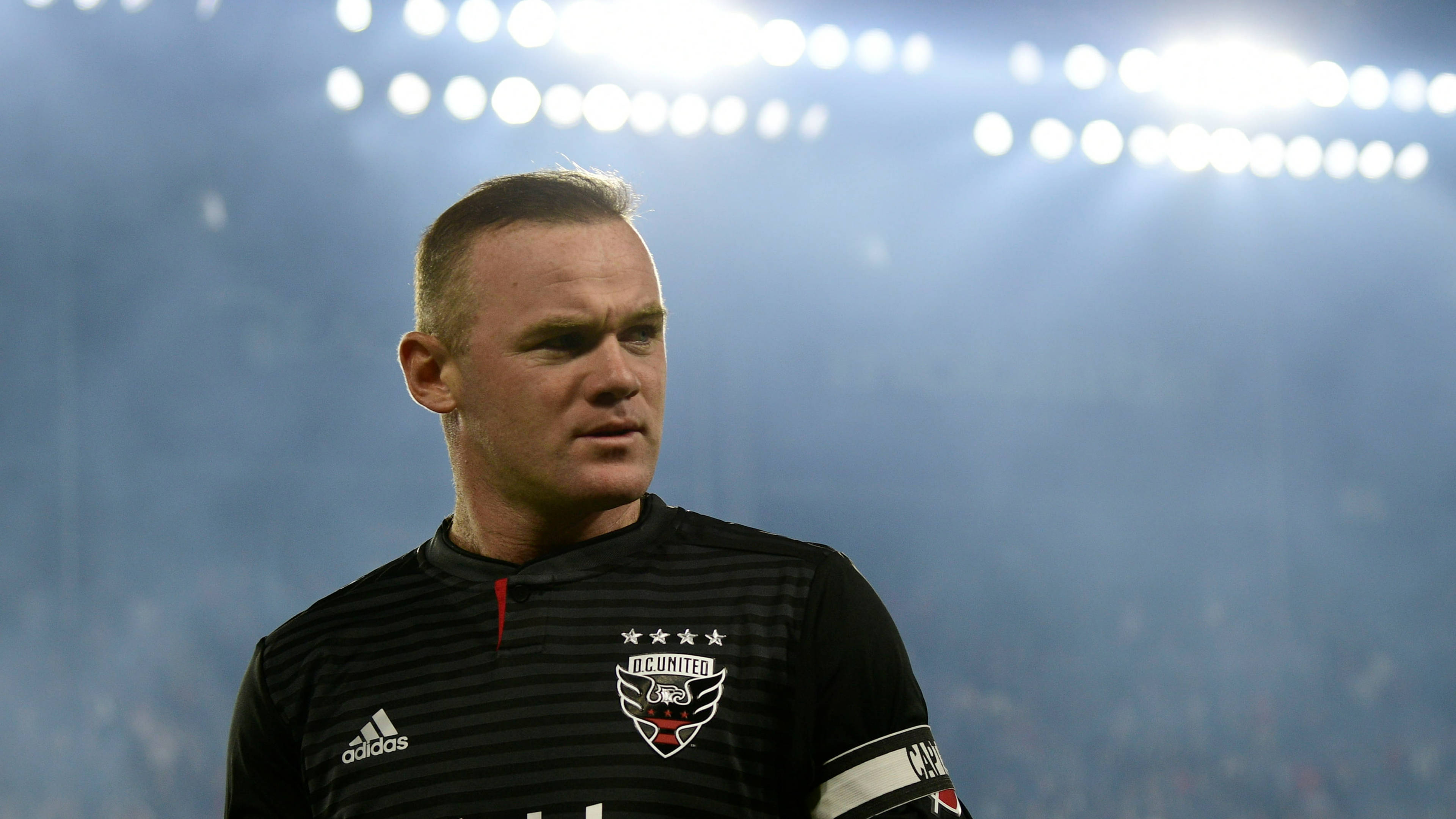 Rooney 桌布 壁紙 — 一位專注的足球員身穿黑色條紋球衣，站在明亮的體育場燈光下，背景是模糊的人群。
