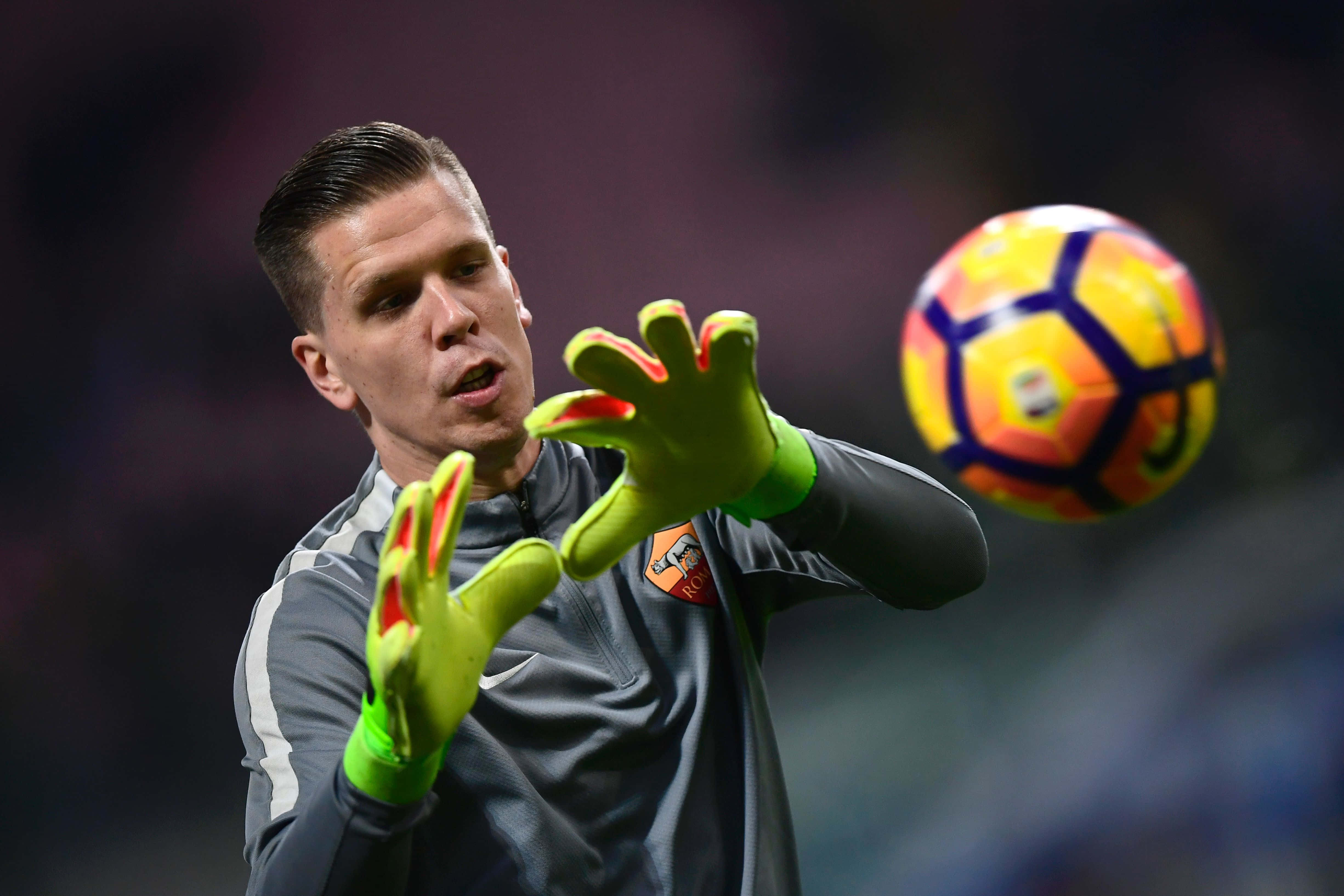Wojciech Szczesny 桌布 壁紙 — 一位專注的足球守門員戴著亮綠色手套，在體育場燈光下於比賽中空中接住一個色彩繽紛的球。